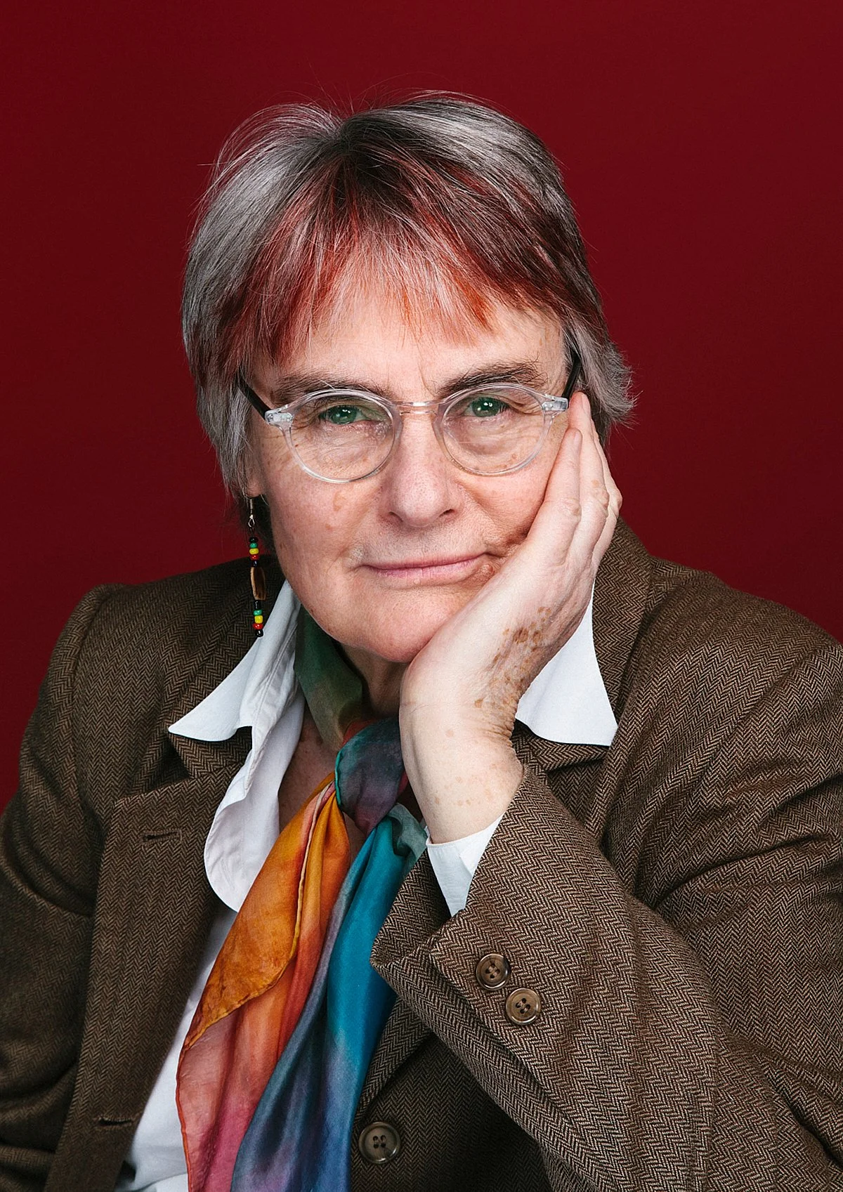 A portrait of an older woman with short gray hair and glasses, resting her face on one hand, wearing a brown blazer, white shirt, and colorful scarf, against a red background.