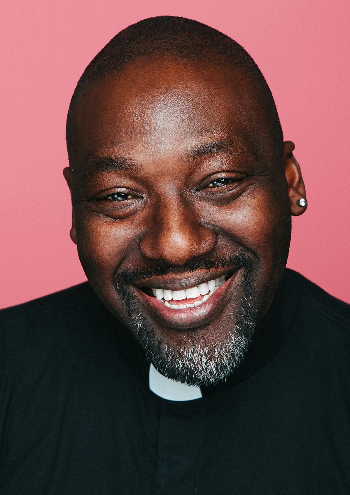 Close-up portrait of a smiling African American man with a beard, wearing a clerical collar and a black suit, against a pink background.