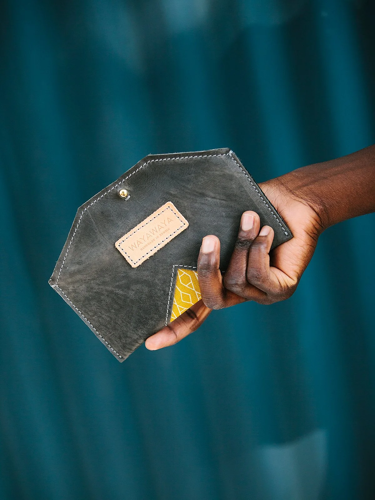 A person holding a dark gray wallet with a cream-colored label that says 'WAWA YAYA'. The wallet also has a small yellow book or card peeking out from a pocket, all against a blurred teal background.