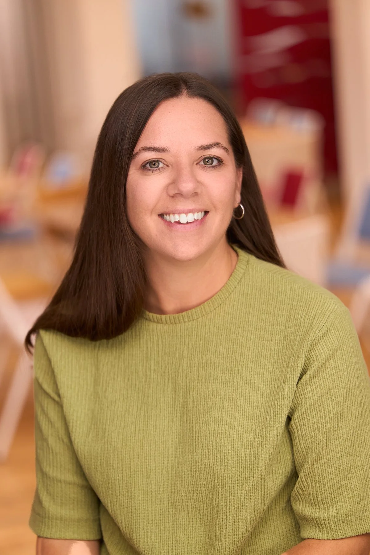 A young woman with long brown hair, green eyes, and light skin, smiling while wearing a green sweater, in an indoor setting with blurred background.