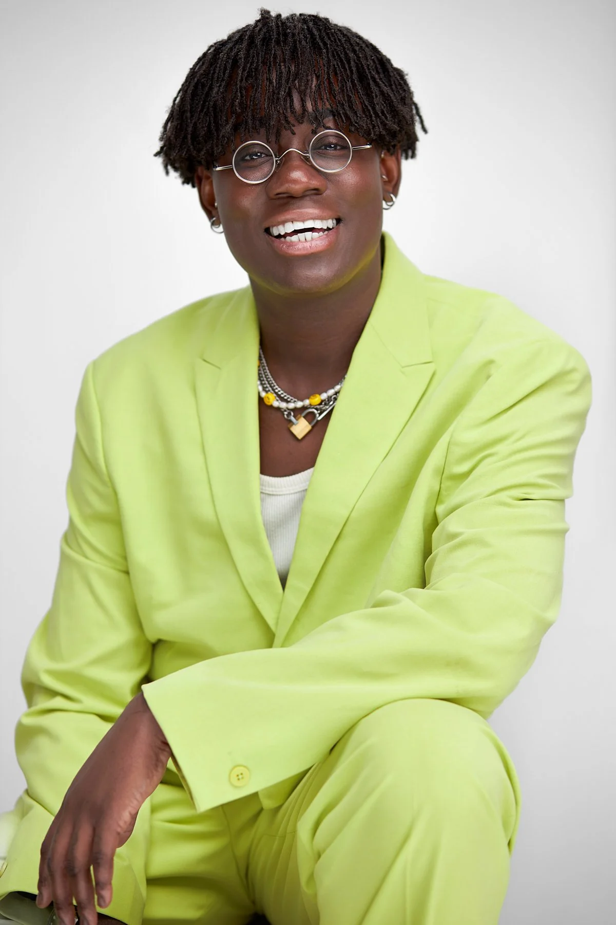 A person with short dreadlocks wearing round glasses, a light green suit, and multiple layered necklaces, sitting against a plain white background, smiling at the camera.