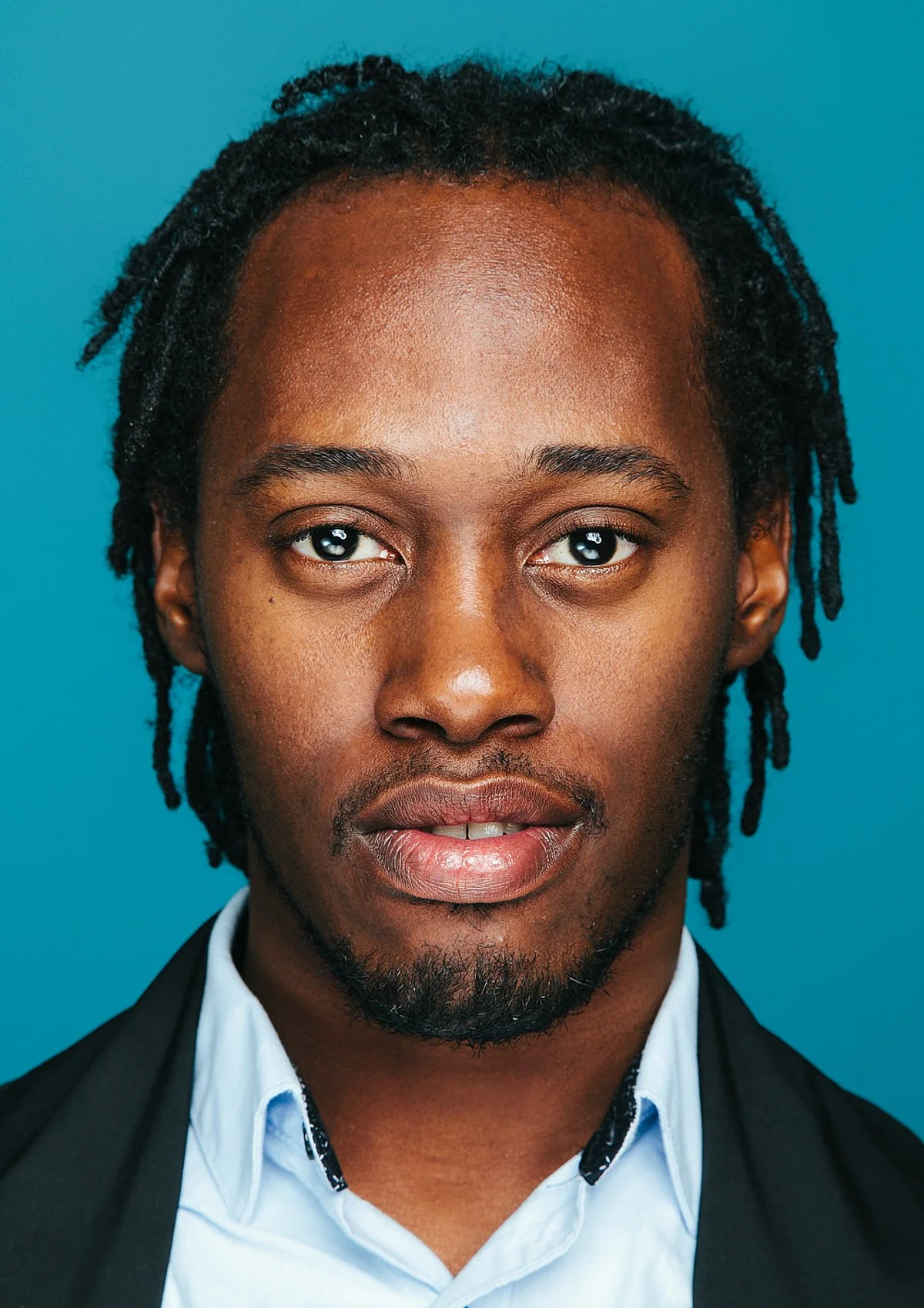 Close-up portrait of a young Black man with dreadlocks, wearing a white shirt and a dark blazer, against a blue background.