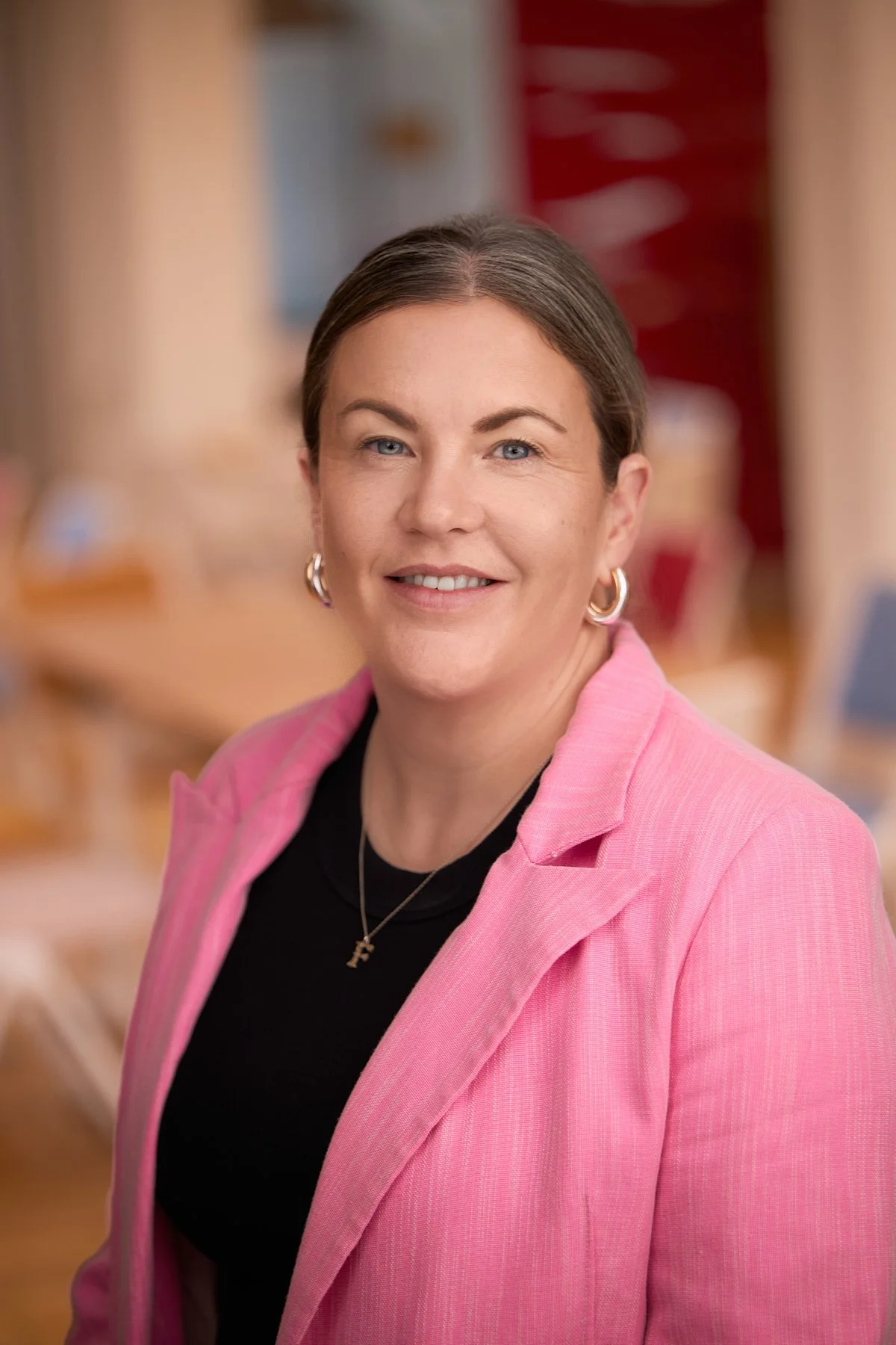 A woman with brown hair, blue eyes, and hoop earrings wearing a pink blazer over a black top, smiling at the camera in an indoor setting.
