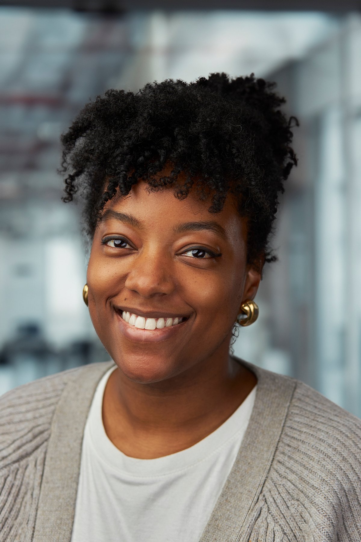 A smiling woman with natural curly hair, wearing gold hoop earrings, a white shirt, and a beige cardigan, in an indoor setting with a blurred background.