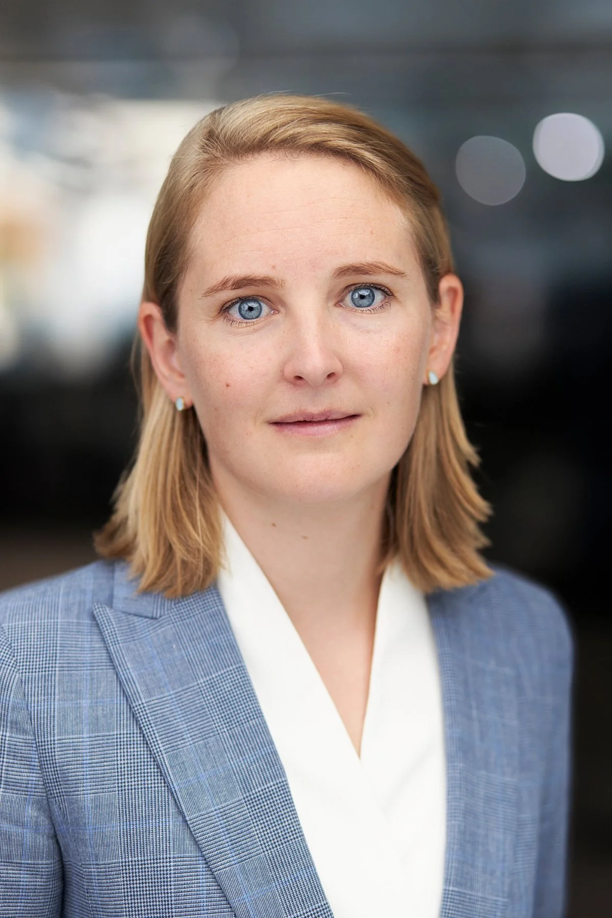 A woman with blonde hair, blue eyes, and light skin wearing a gray plaid blazer and a white shirt, standing outdoors with blurred city lights in the background.