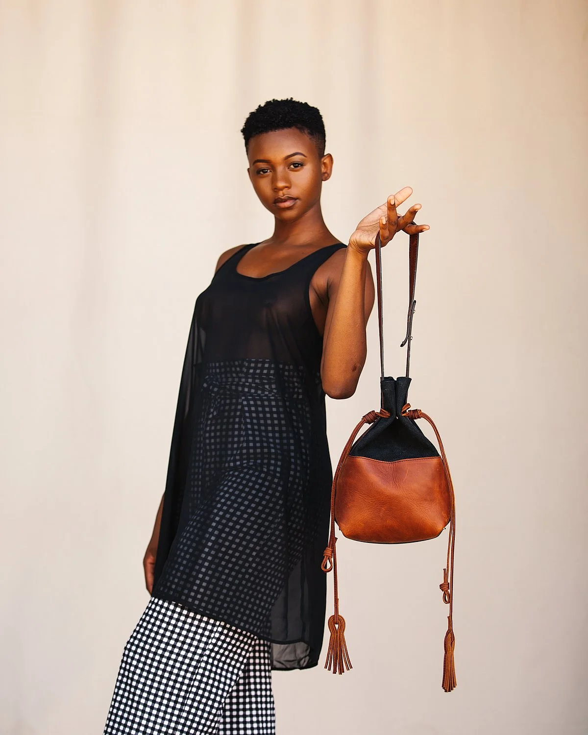 A young woman with short curly hair wearing a sheer black top and black-and-white checkered pants, holding a brown and black handbag, standing against a plain background.