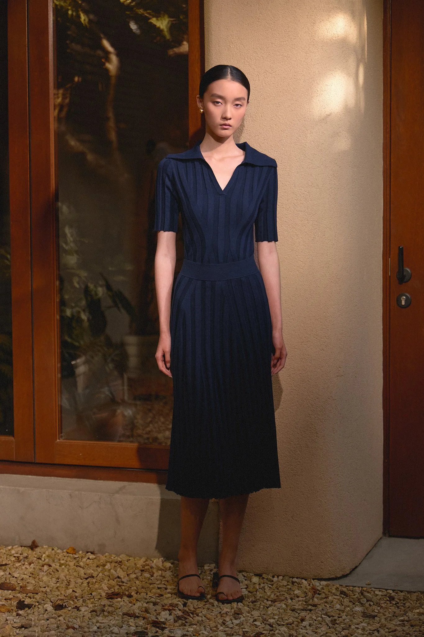 Fashion lookbook: model in a navy pleated dress by a glass door