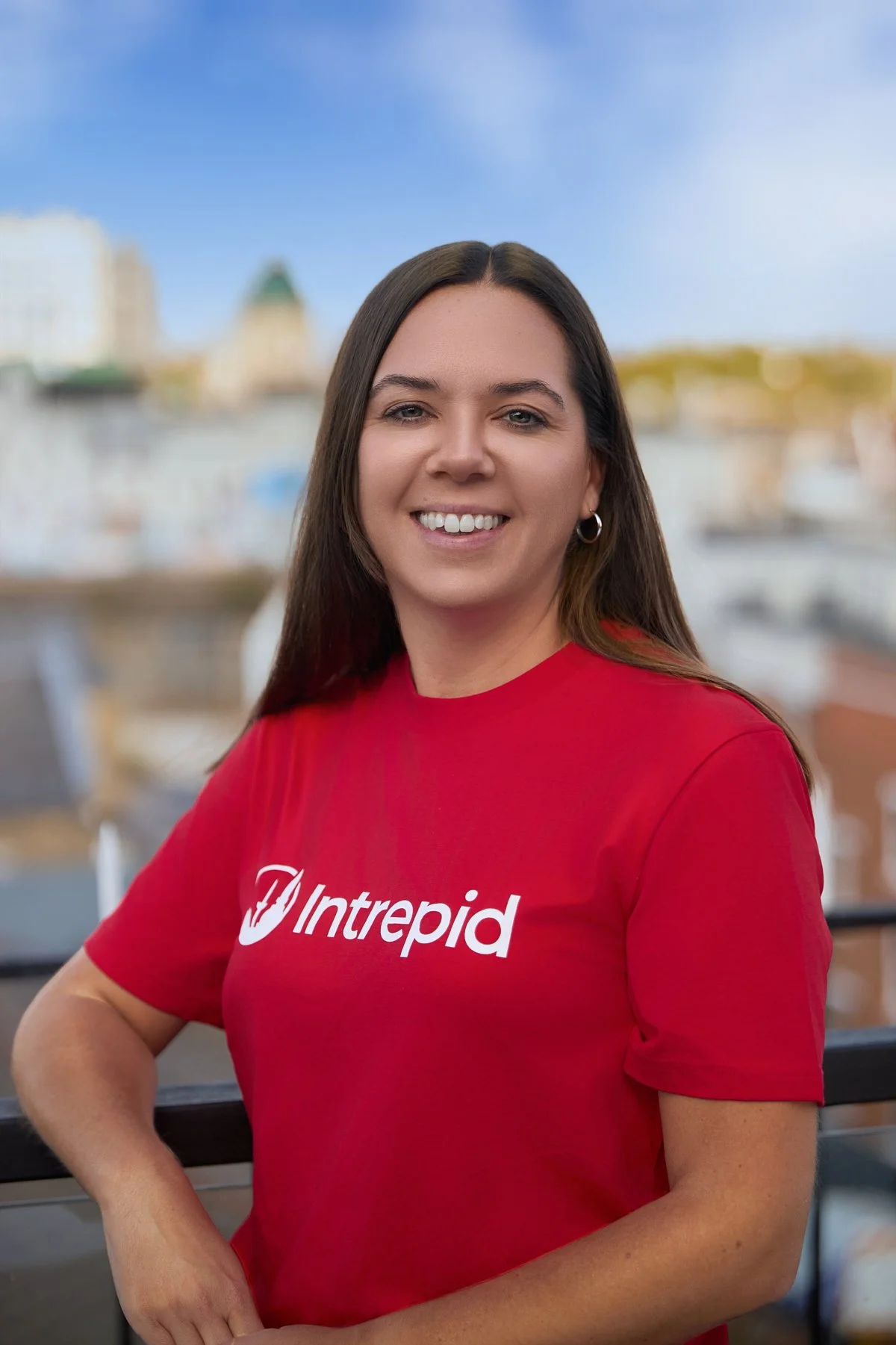 A woman with long brown hair, wearing a red T-shirt with the logo 'Intrepid', smiling, with water and city buildings in the background during daytime.