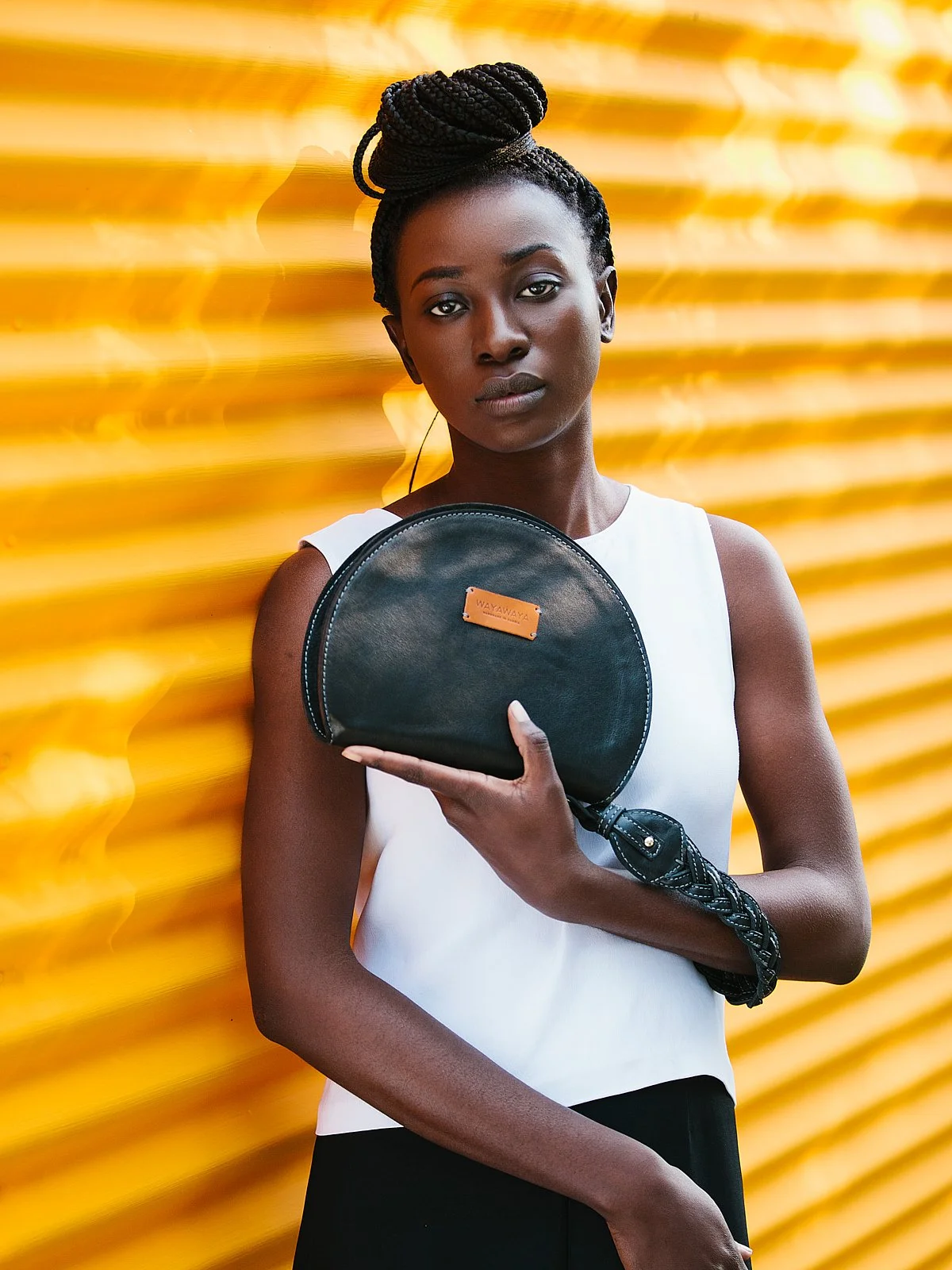 A woman with dark skin and braided hair styled in an updo, wearing a white sleeveless top, holding a black semi-circle purse with a braided strap, standing against a yellow corrugated metal background.