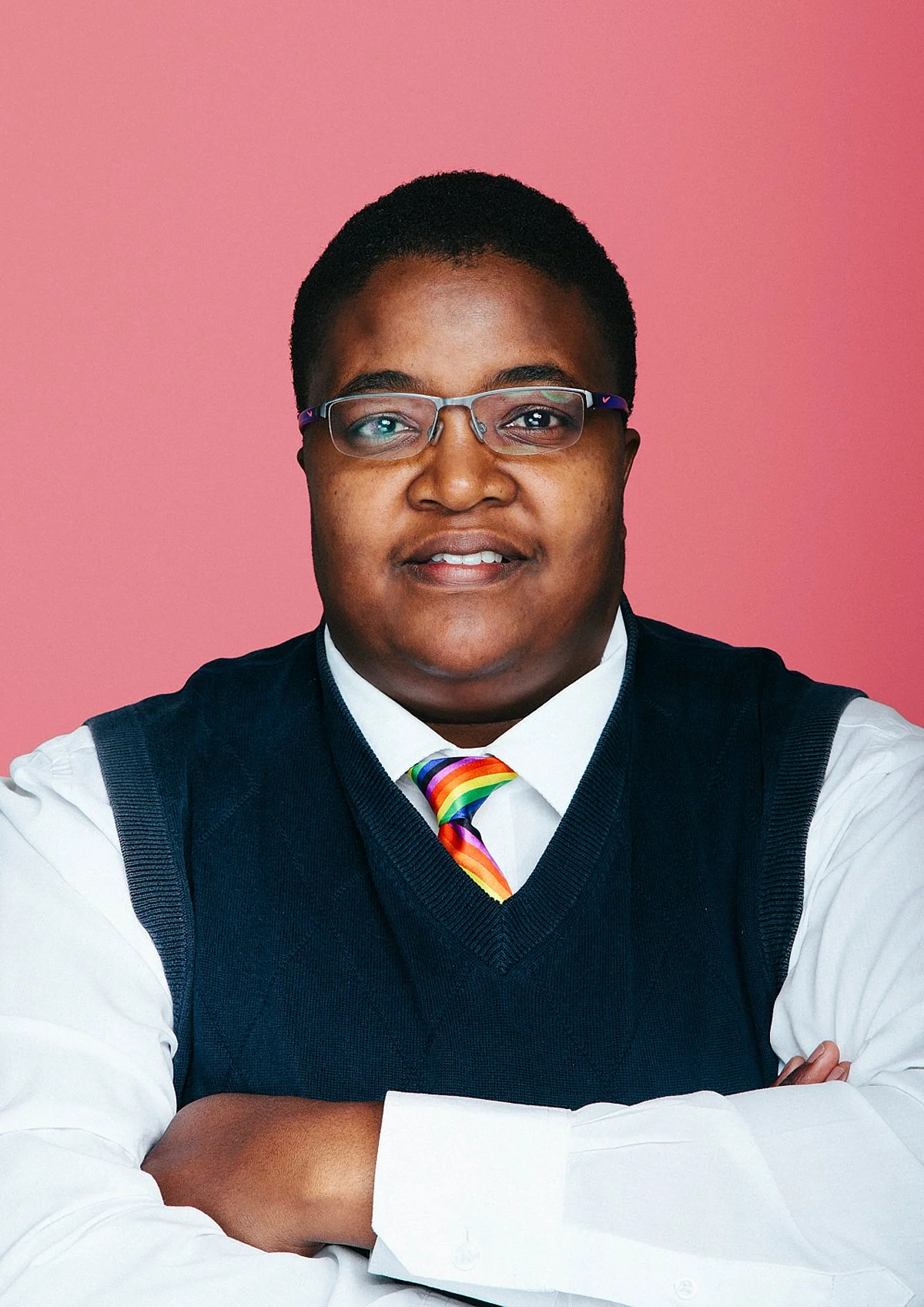 Portrait of a person with short hair, glasses, wearing a white shirt, a rainbow-striped tie, and a navy blue vest, standing against a pink background.