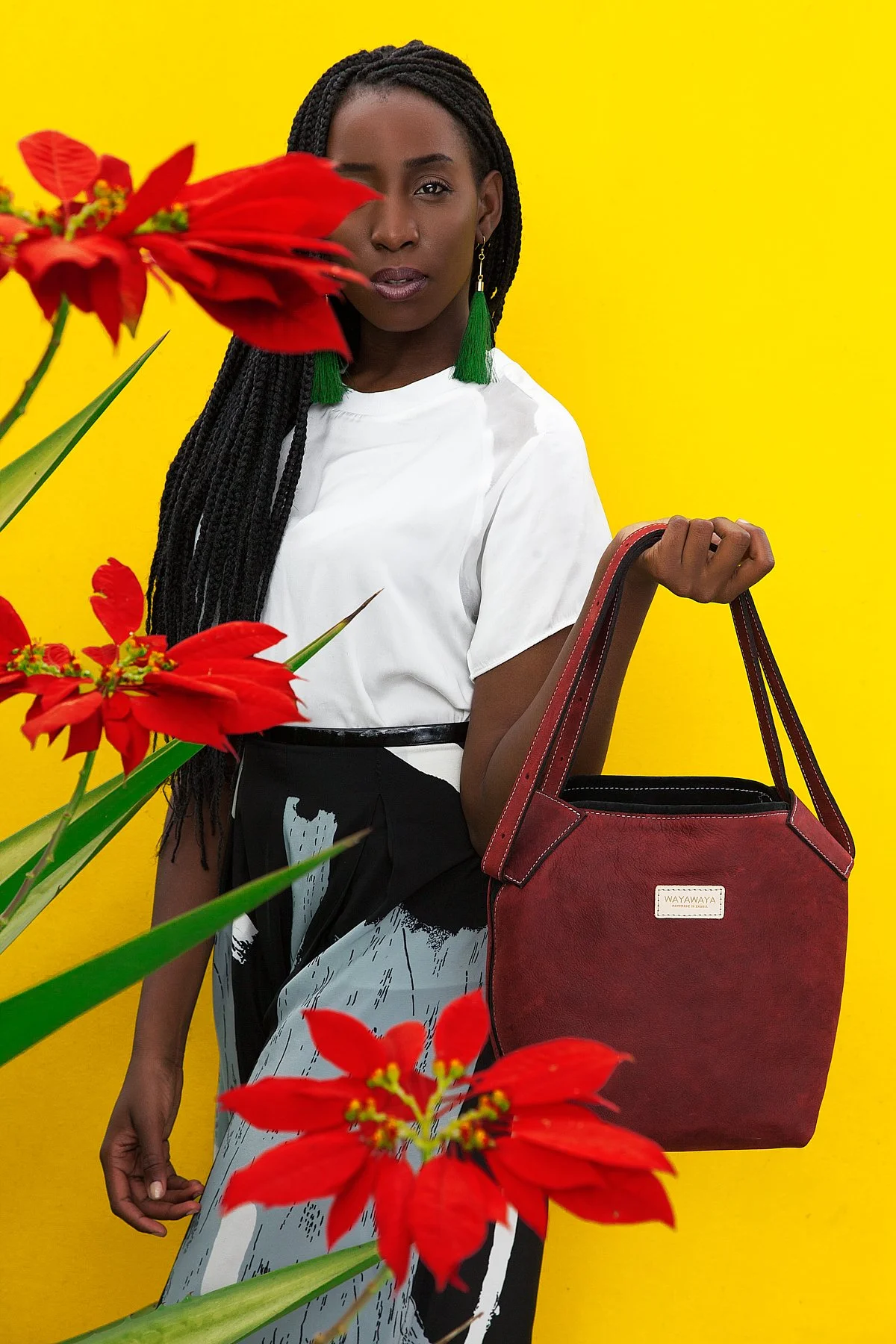 A woman with dark skin and long braided hair stands against a bright yellow background, partly covered by red flowers in the foreground, holding a red handbag and wearing a white blouse with green tassel earrings.