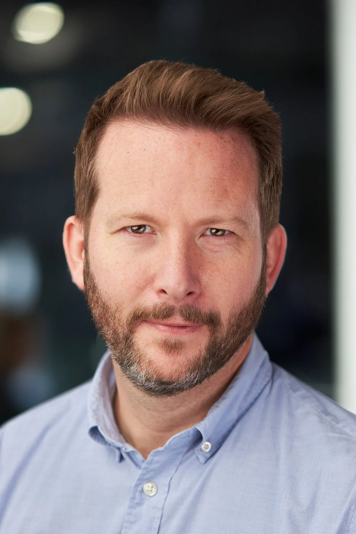 Close-up of a man with short brown hair, beard, wearing a light blue button-up shirt.