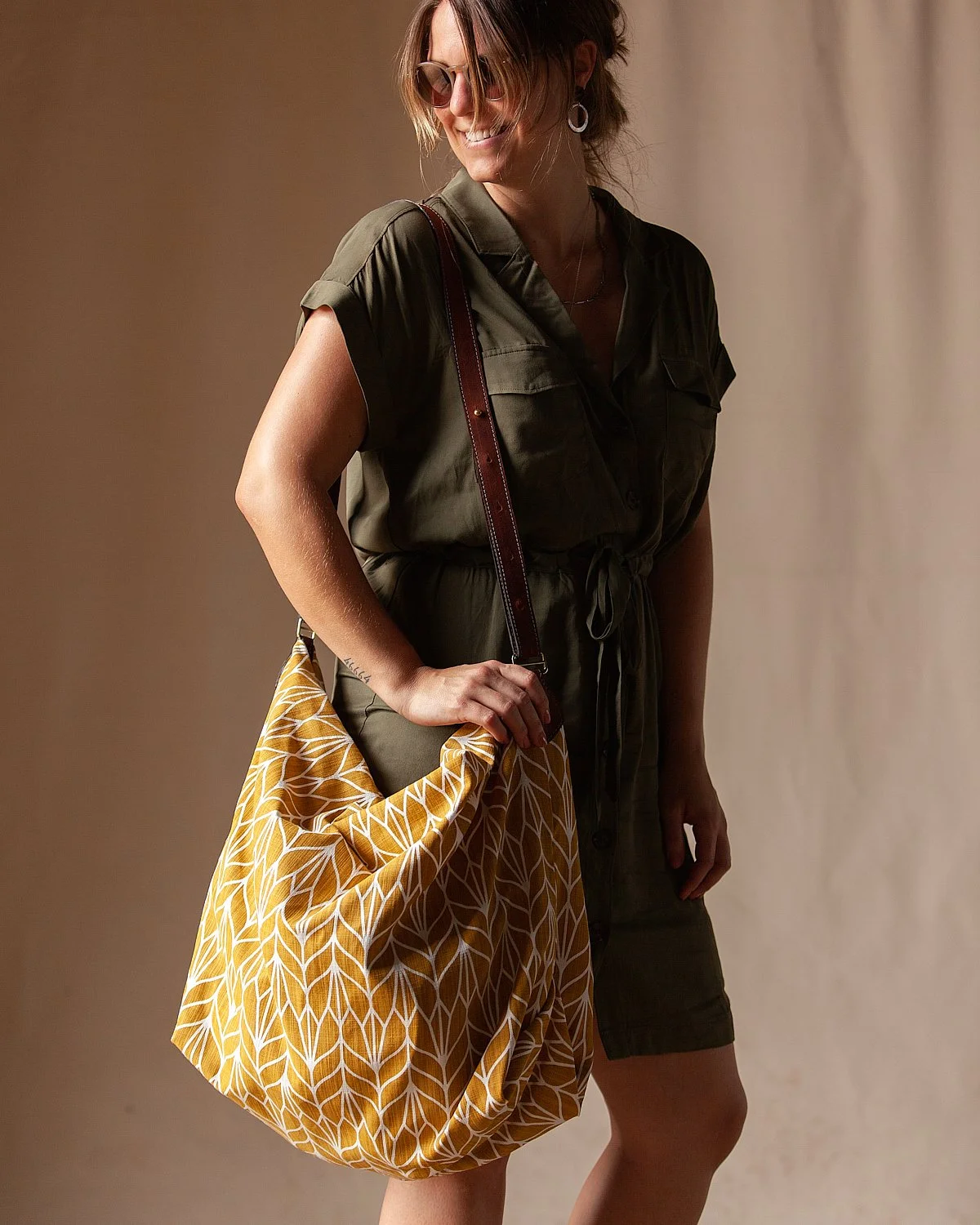 A woman holding a yellow patterned bag, wearing sunglasses, a dark green short-sleeved jumpsuit, and earrings, standing against a beige background.