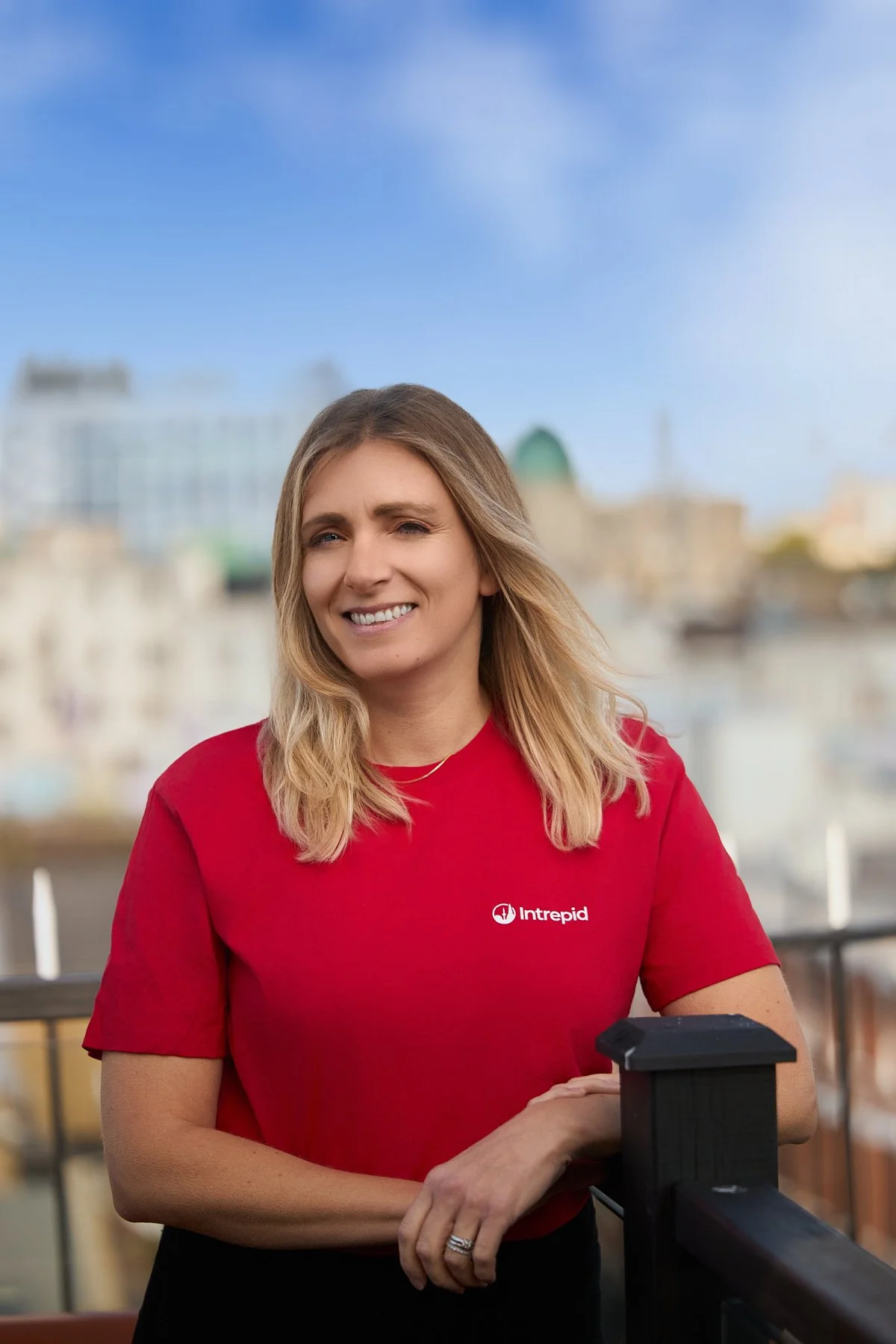 A woman smiling outdoors on a balcony, wearing a red shirt with the Intrepid logo.