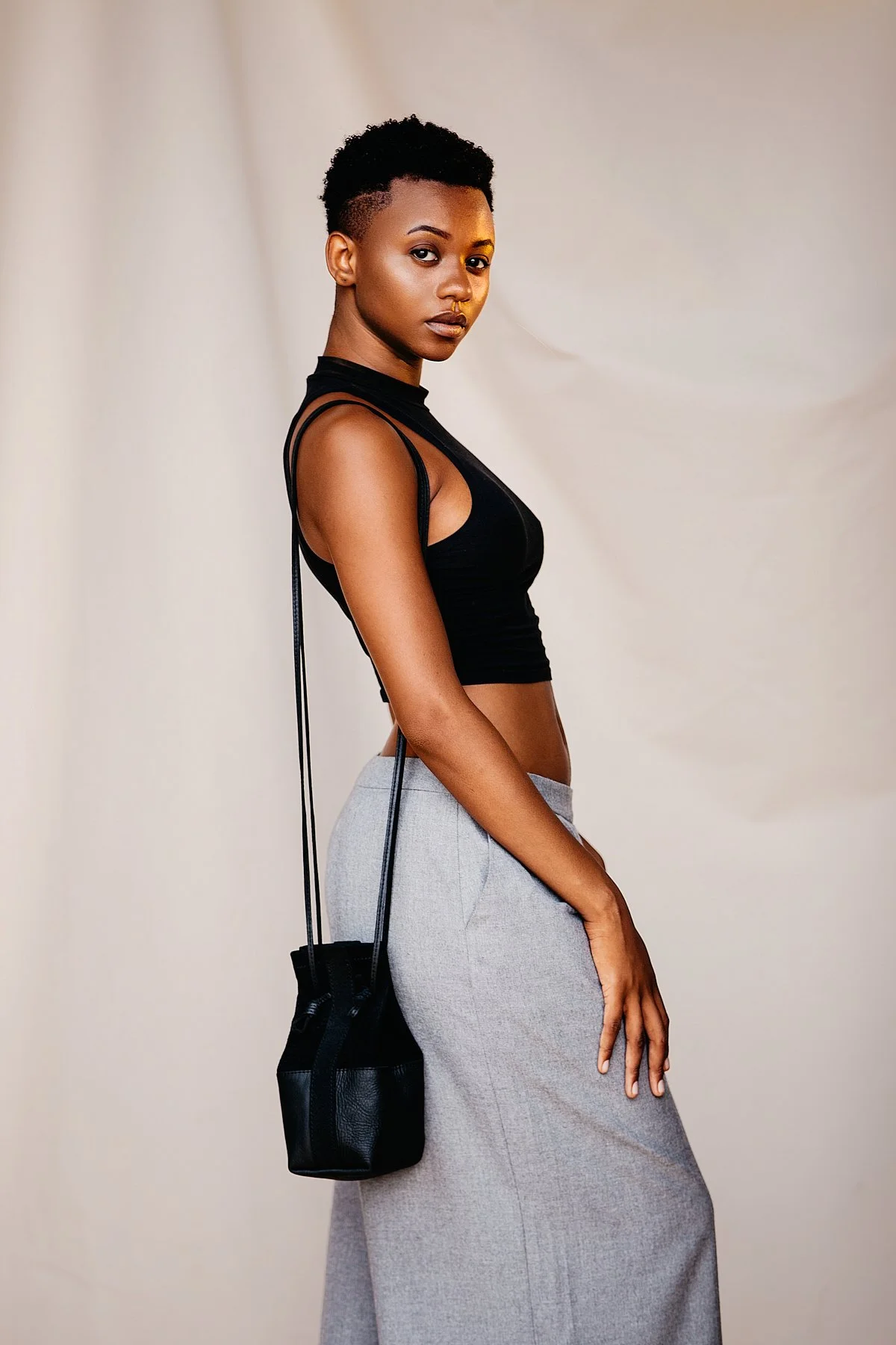 A woman with dark skin and short curly hair, wearing a black sleeveless top and a gray skirt, standing against a plain neutral background, with a black bag hanging from her shoulder.