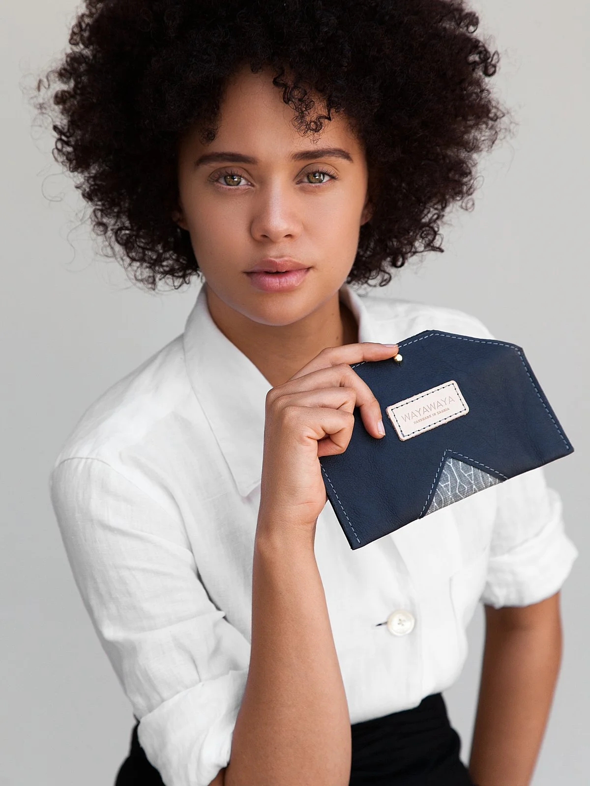 Young woman with curly hair wearing a white button-up shirt, holding a dark blue wallet with a brand label, against a plain light background.