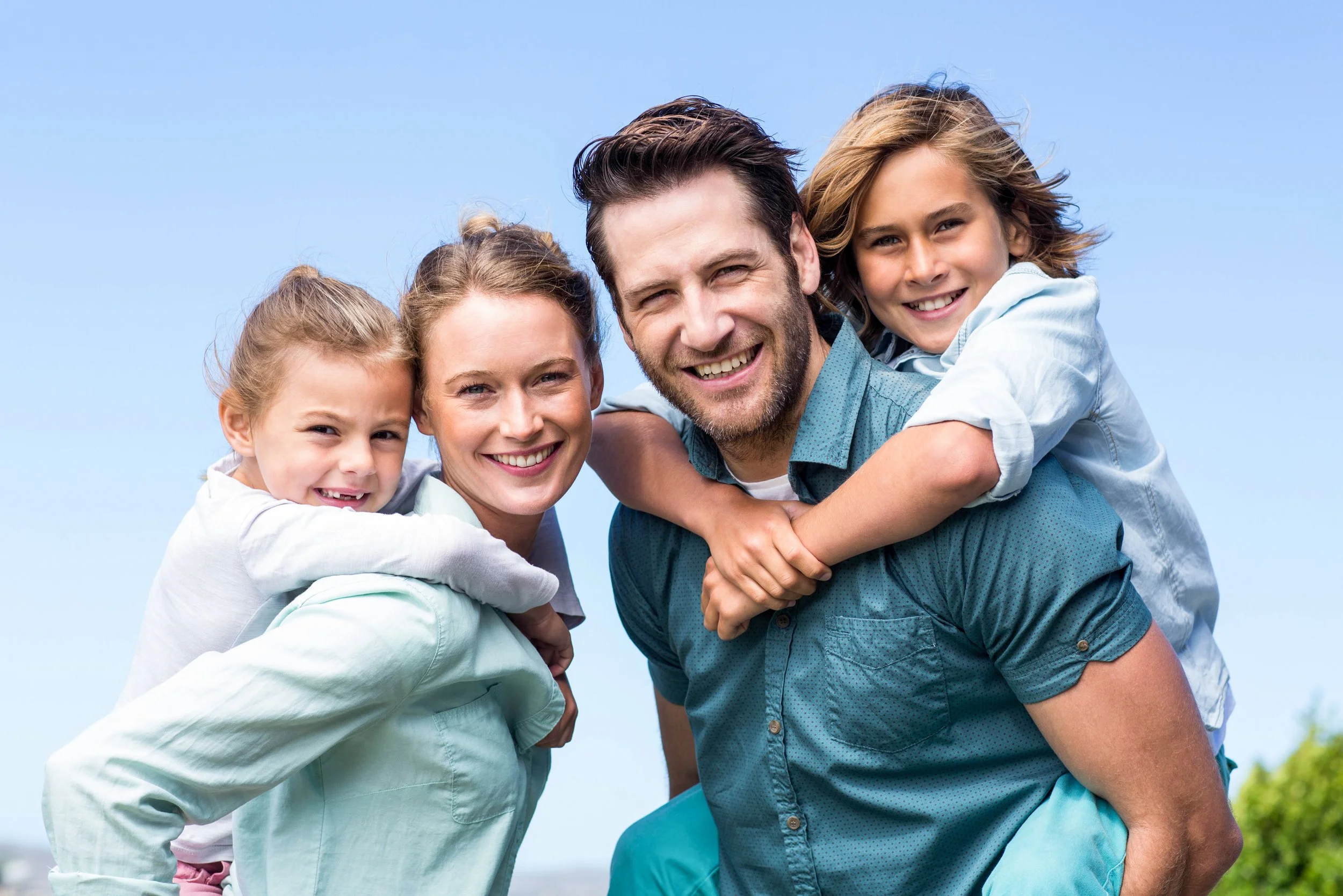 A happy family of five, including two young girls, a woman, a man, and a teenager, outdoors under a clear blue sky.