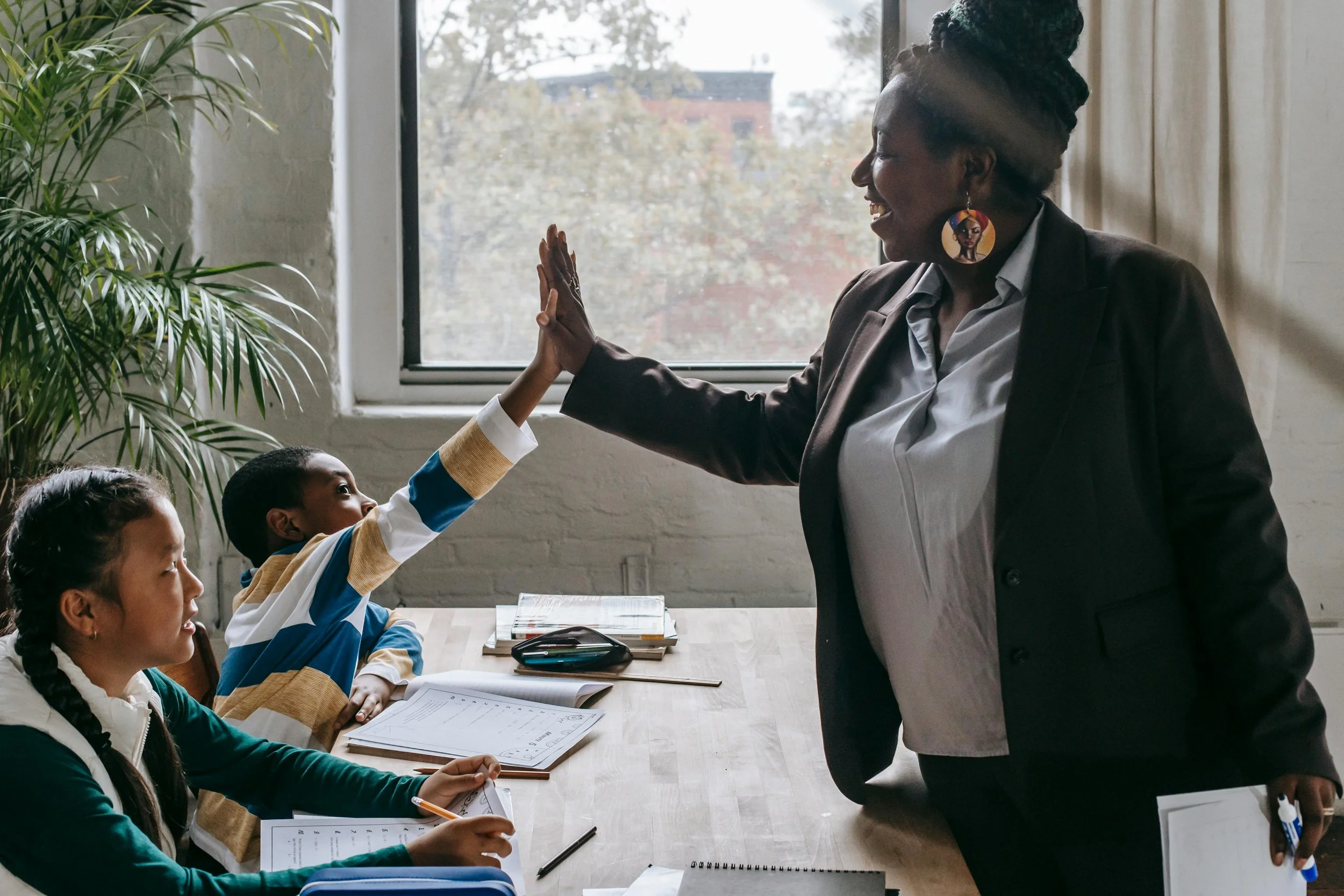 A photo of a teacher in a classroom wearing a dark blazer highfiving a child in a striped shirt.