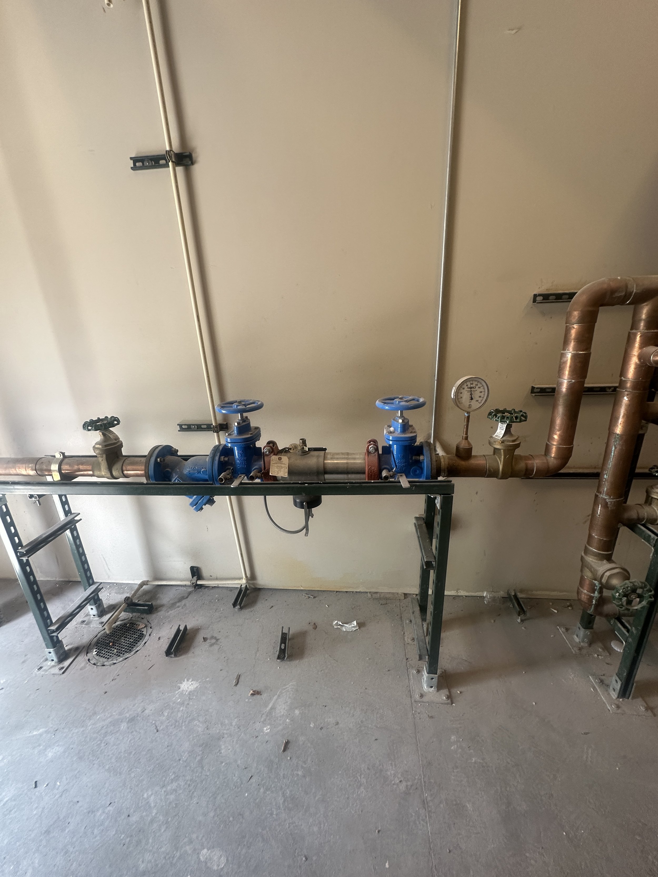 Industrial water pipes and valves installed on a metal frame against a beige wall, with a pressure gauge and various fittings.