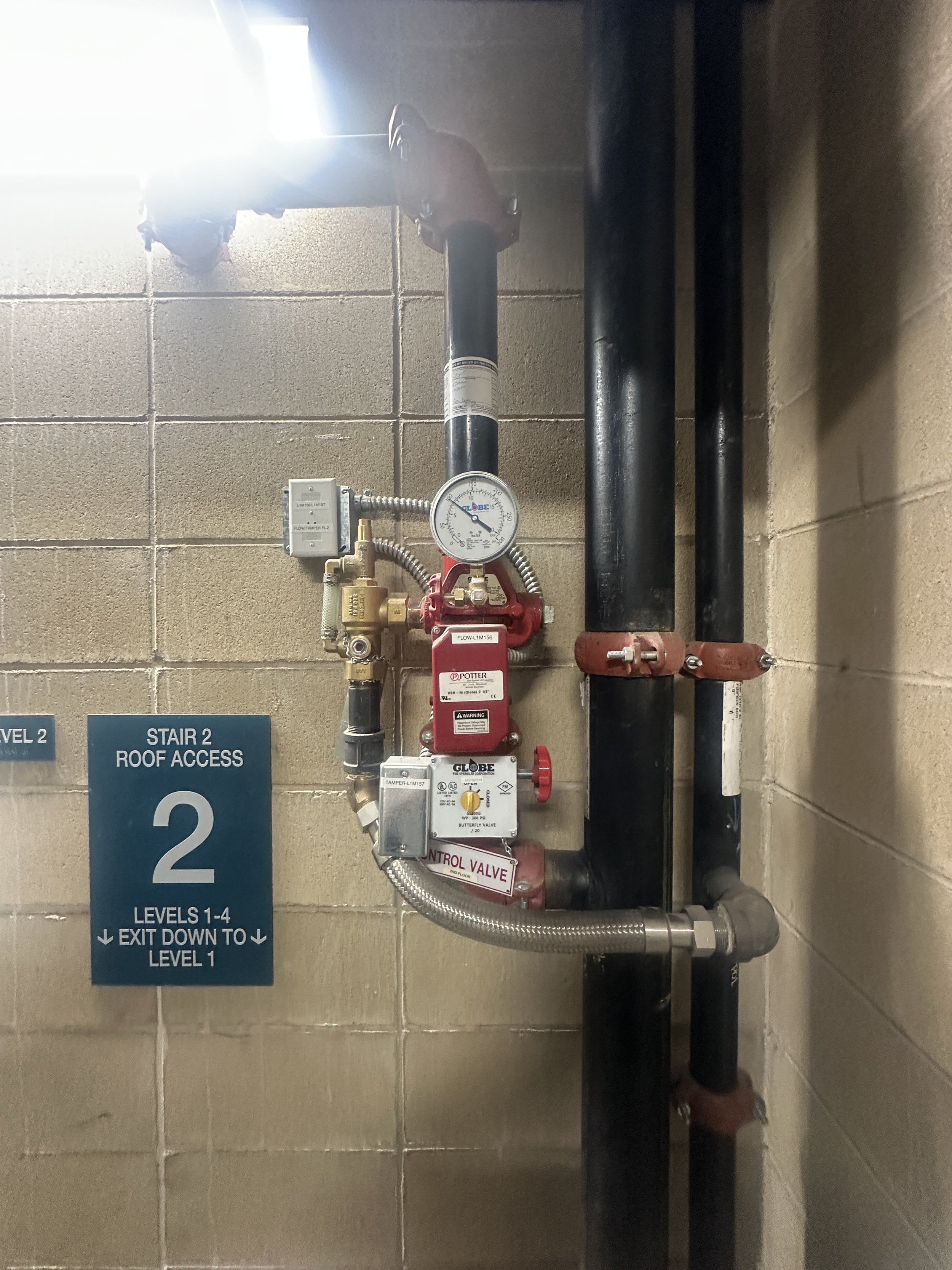 Mechanical piping system with gauges, valves, and pipes mounted on a beige brick wall near a stairway sign.