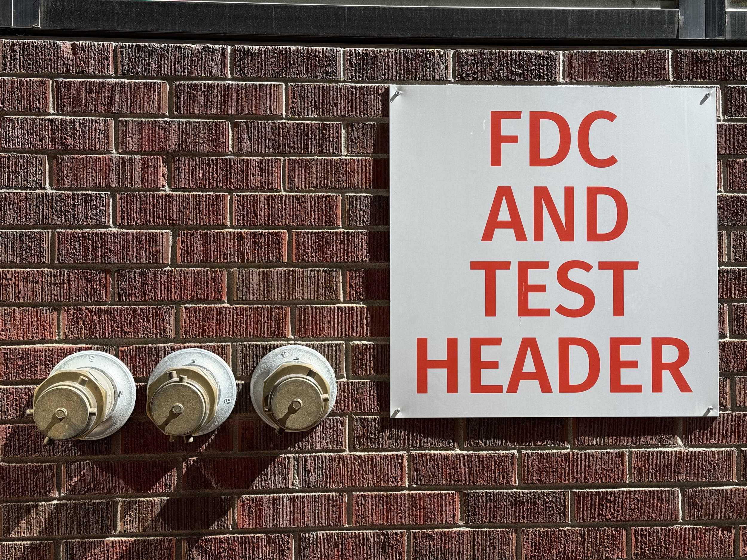 Brick wall with a white sign reading 'FDC AND TEST HEADER' in red letters and three electrical meters beneath the sign.