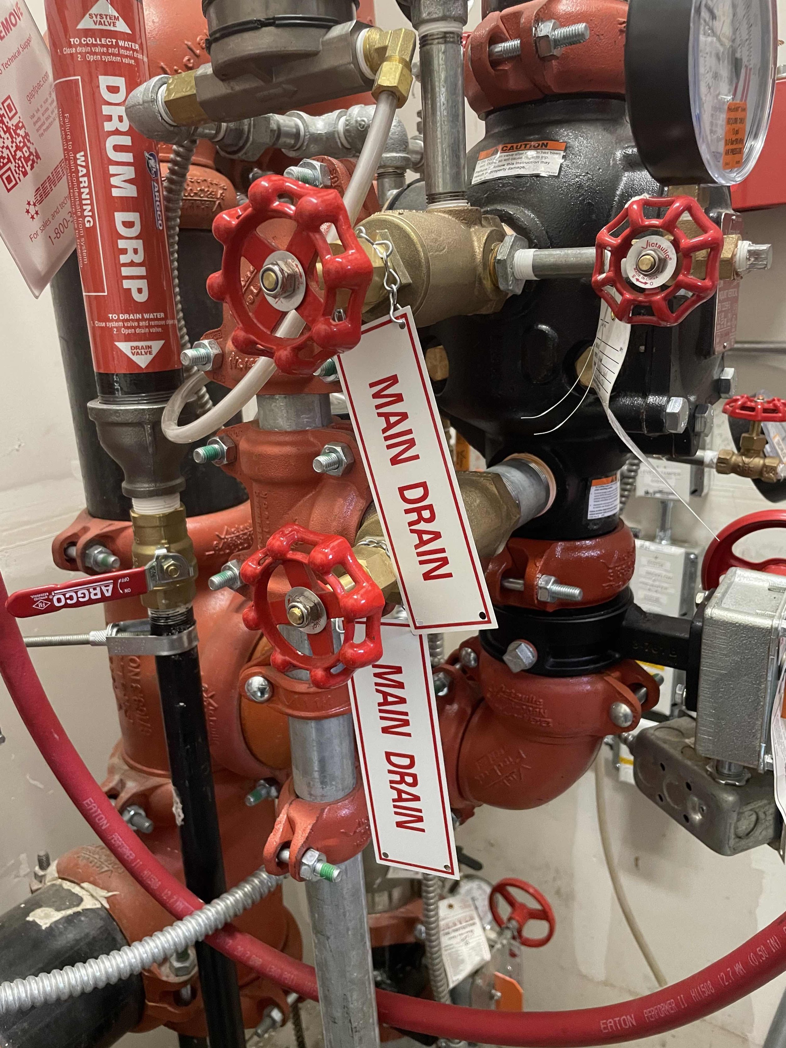 A complex fire protection system with red valves, pipes, and gauges, including a main drain valve with a "MAIN DRAIN" tag, fire sprinkler components, and various fittings.