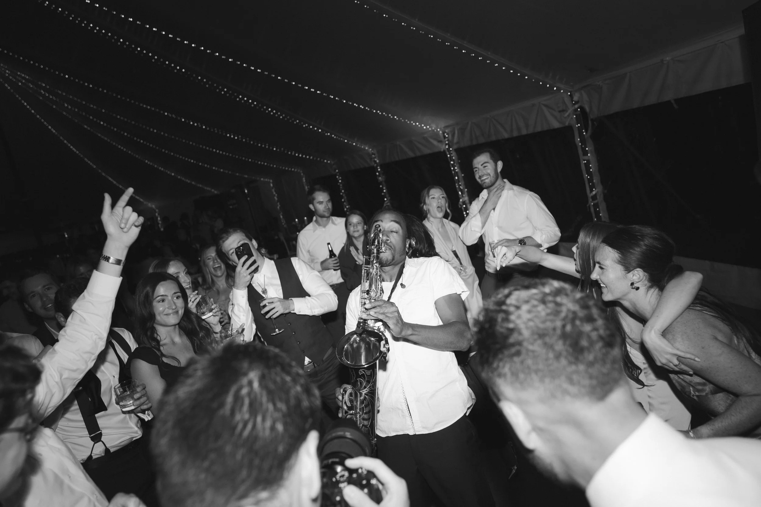 A lively party scene with people dancing and singing, a woman playing a saxophone with others enjoying and taking photos, under string lights in a tent.