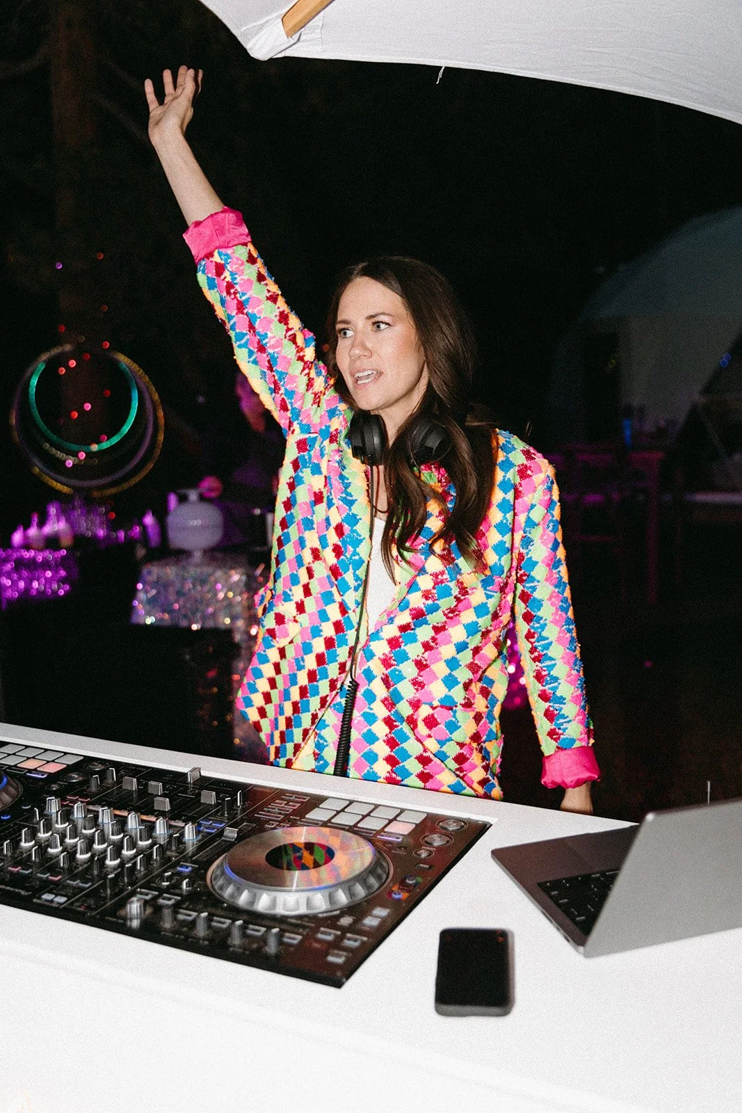 A woman with long brown hair standing behind a DJ table with equipment, wearing a colorful geometric patterned jacket and a white top, raising her right hand and wearing headphones around her neck, in a nightclub or party setting.