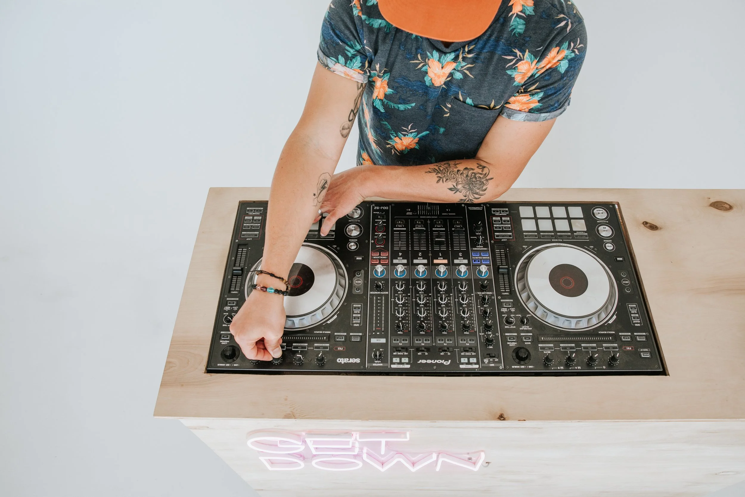 Person wearing a floral shirt and an orange cap DJing with a Pioneer DJ controller on a light wooden table.