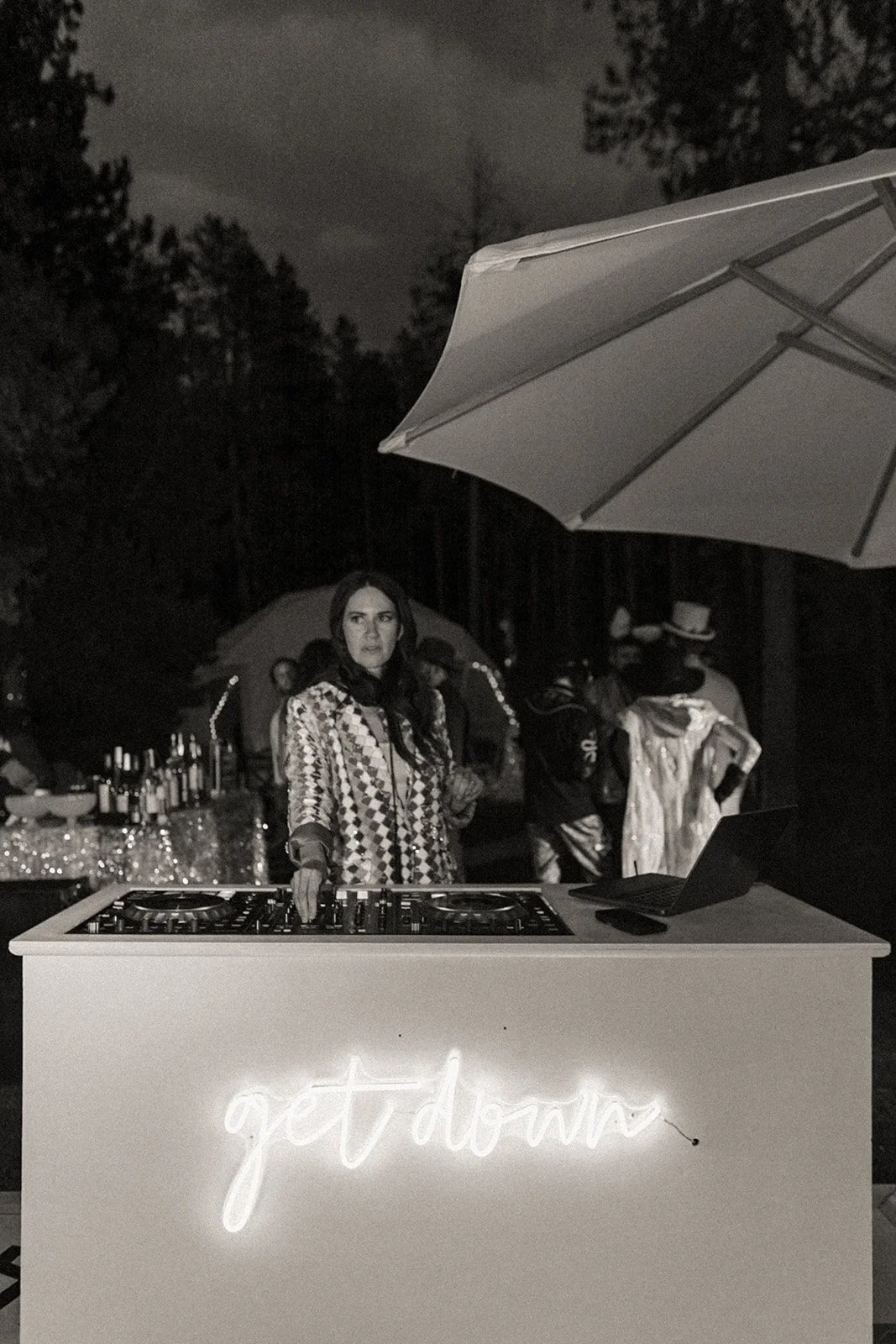 A woman DJ standing behind a DJ booth with a neon sign that says 'get down', at an outdoor nighttime event with trees in the background.