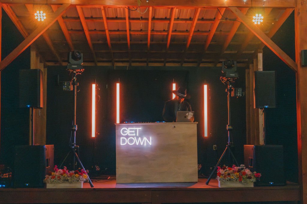 A DJ performing on a stage with a wooden backdrop, illuminated by red vertical lights, with speakers and floral arrangements at the front. A neon sign reads 'GET DOWN'.
