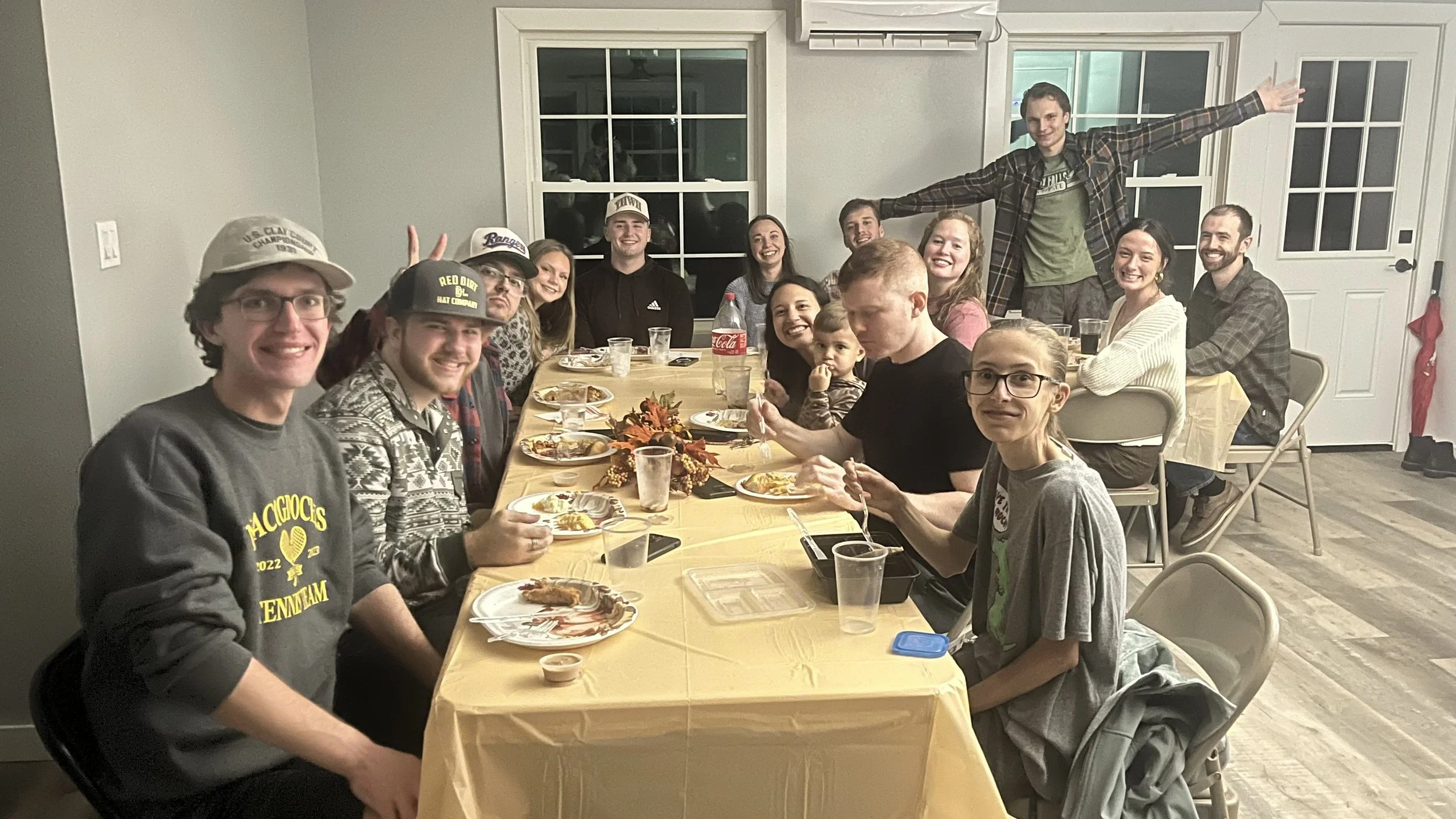 A large group of young adults sitting around a dining table celebrating, with some standing behind, inside a well-lit room with white walls and windows, enjoying a Friendsgiving meal and smiling.