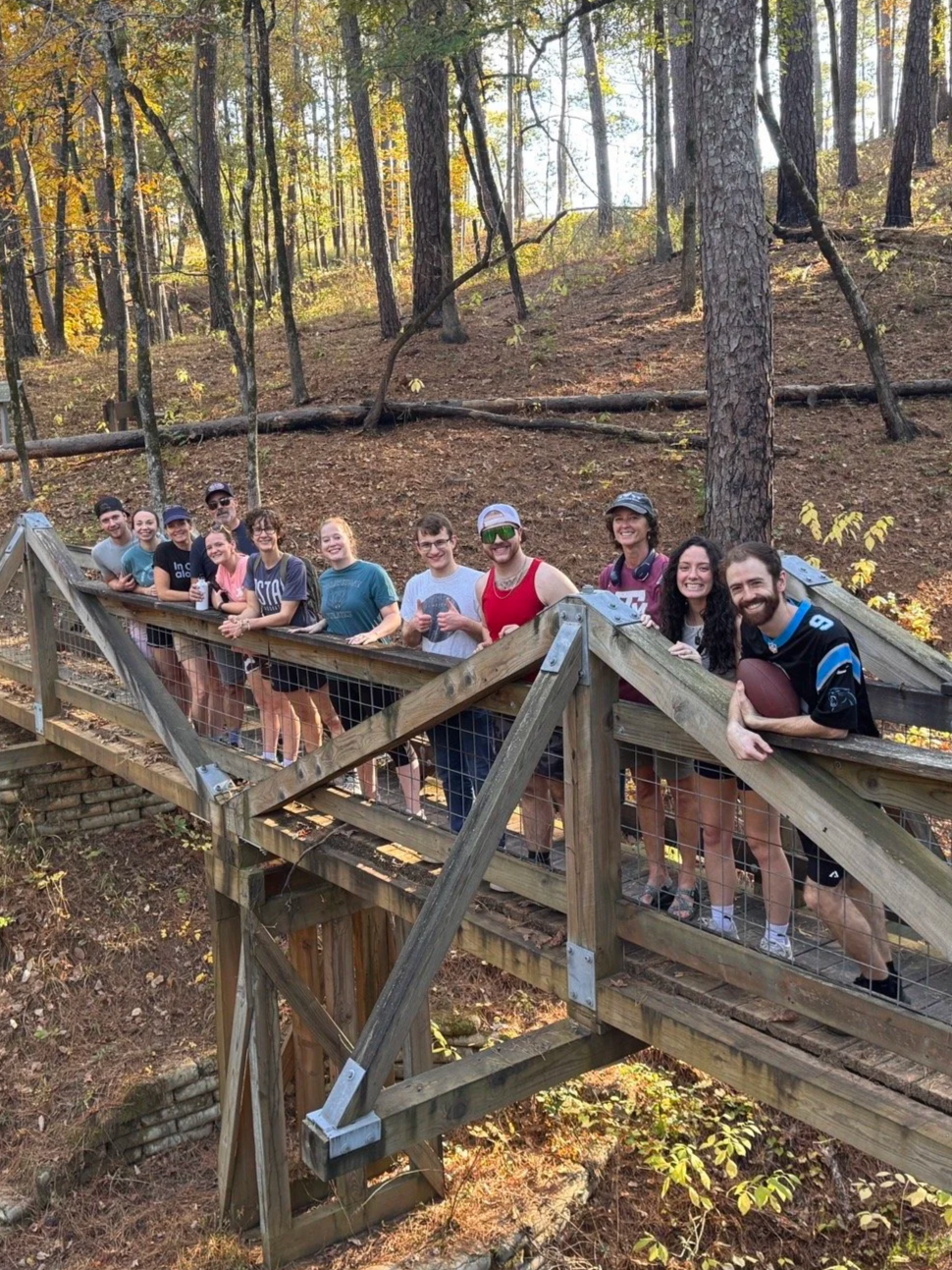 A group of eleven young adults standing on a wooded outdoor bridge, smiling at the camera while hiking.
