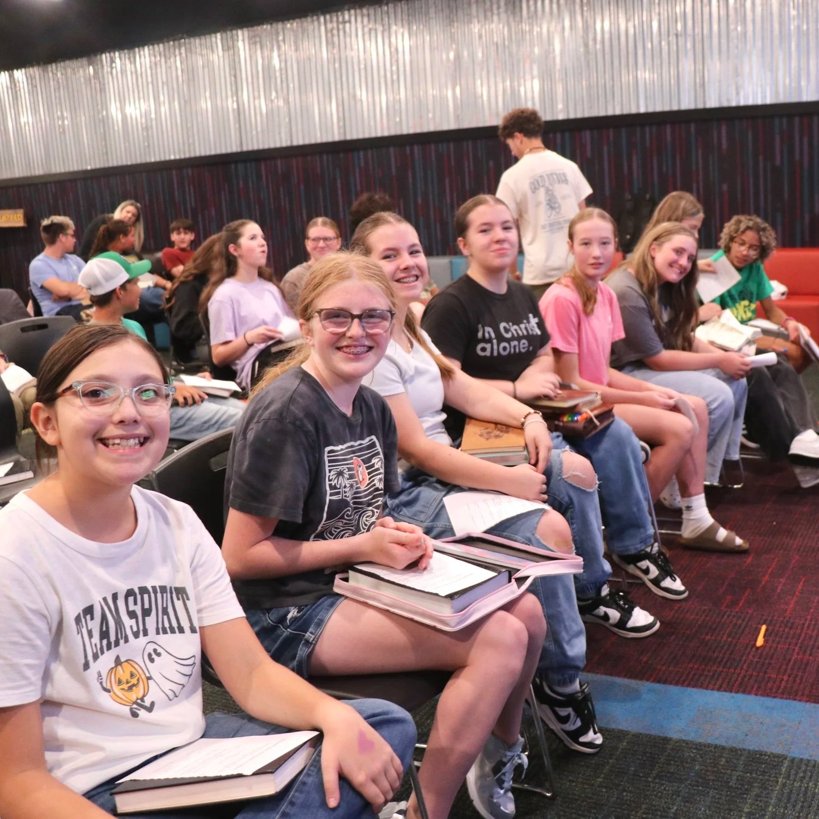 Group of smiling children sitting in the Youth House ready for the sermon, holding notebooks and papers, some looking towards the camera.