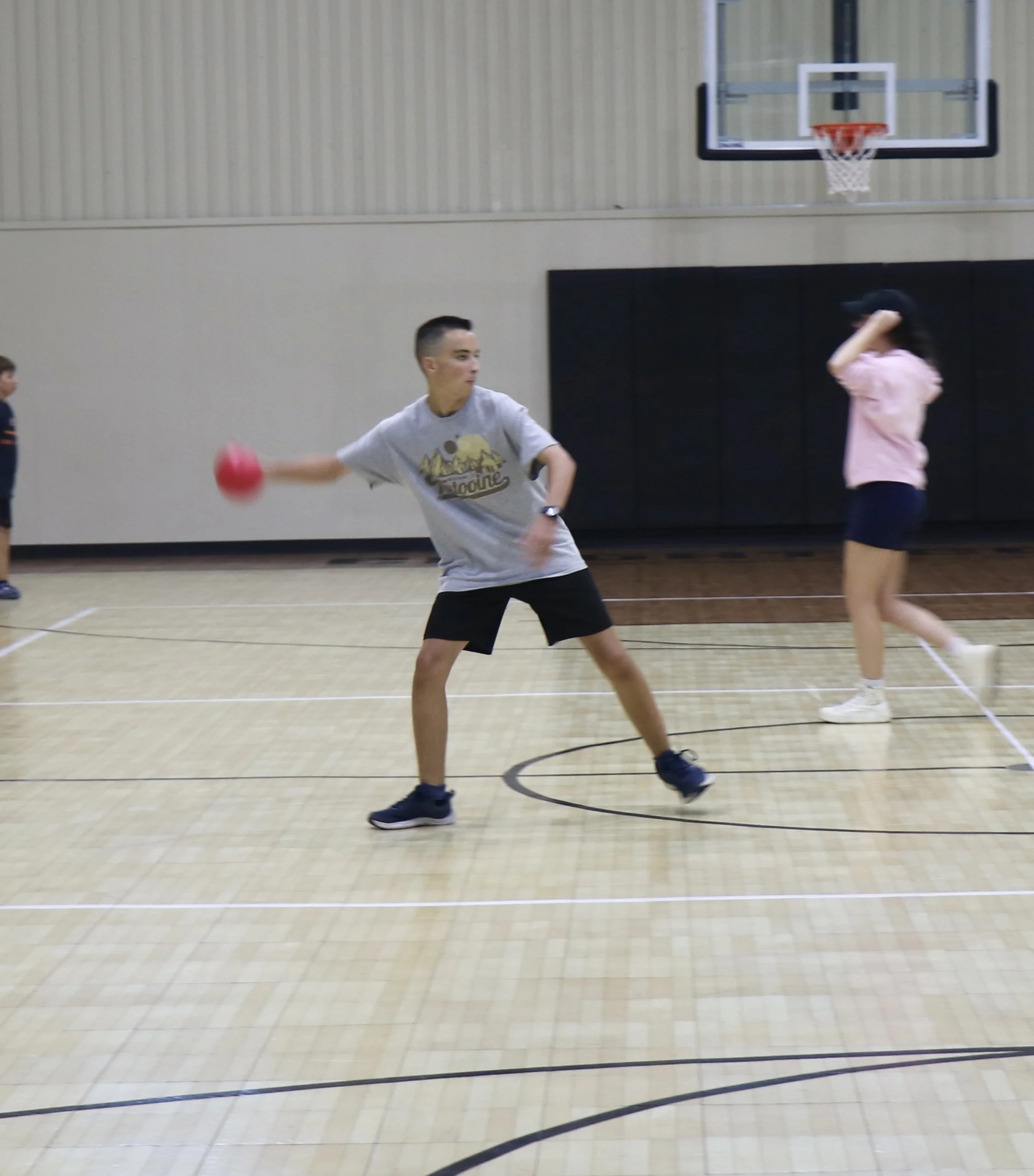 Young man playing dodgeball in a gymnasium, with a basketball hoop in the background.