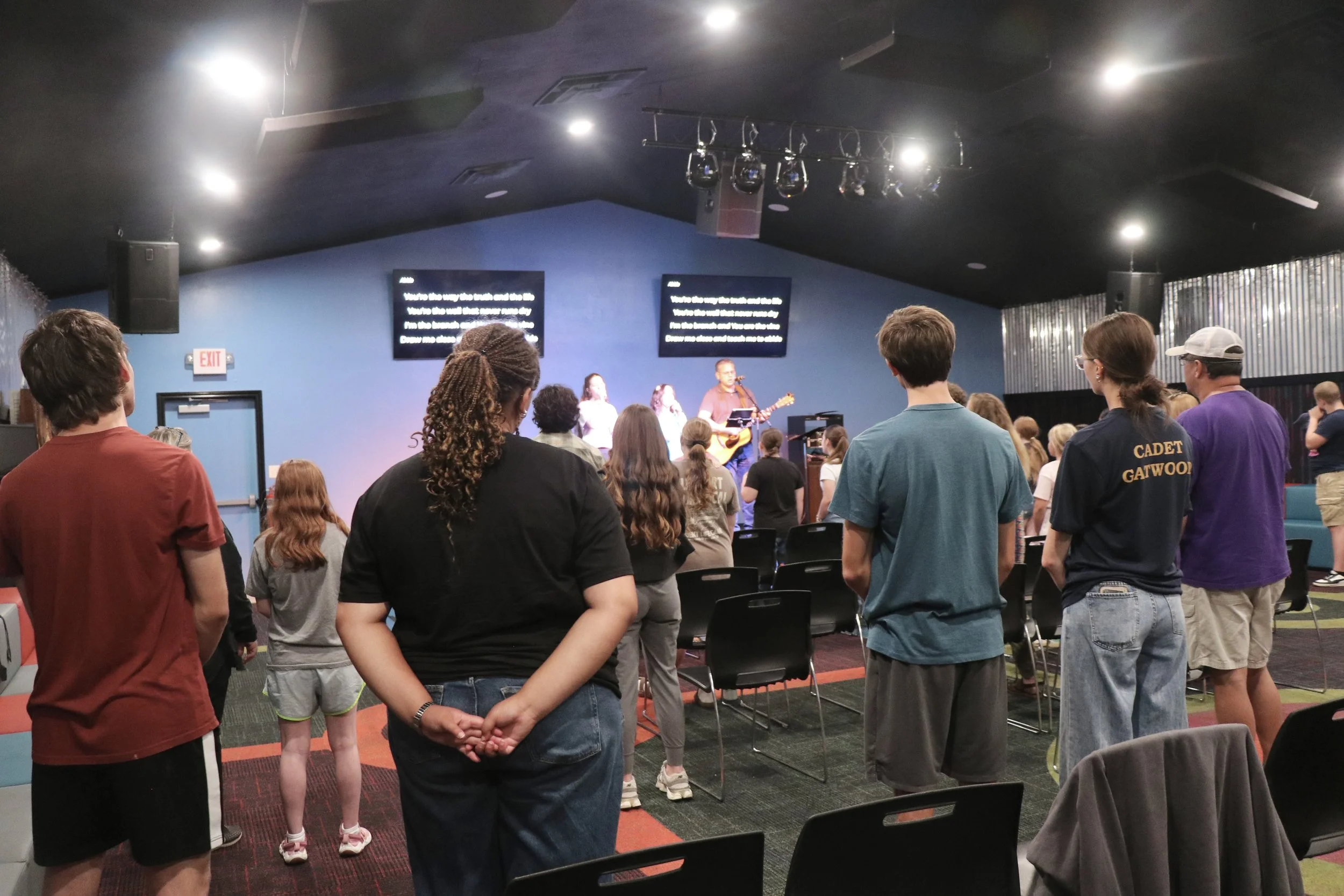 A group of people standing and facing a stage where three youth are leading worship, one playing guitar, with lyrics displayed on screens behind them in a dark indoor setting.