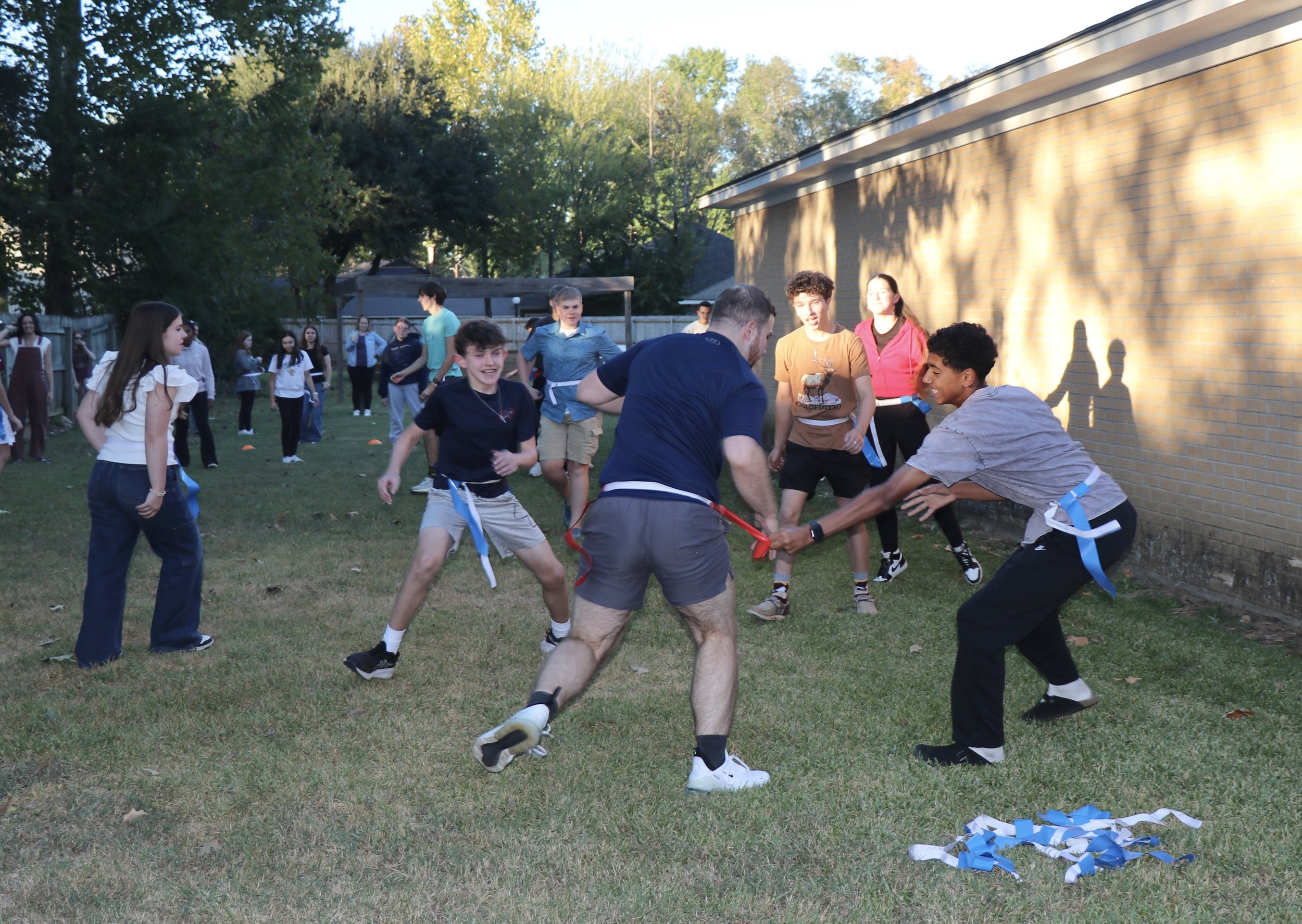 A group of kids and teens playing capture the flag outdoors on a grassy yard with trees and a brick building in the background, one pulling the flag while others cheer and watch.