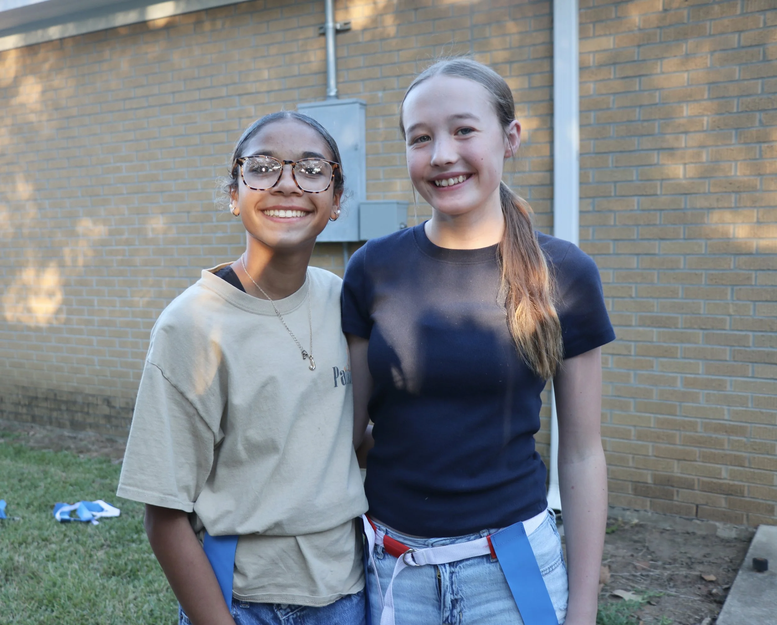 Two girls smiling outdoors near a brick wall, one wearing glasses and a beige t-shirt and the other with a black t-shirt and long hair.