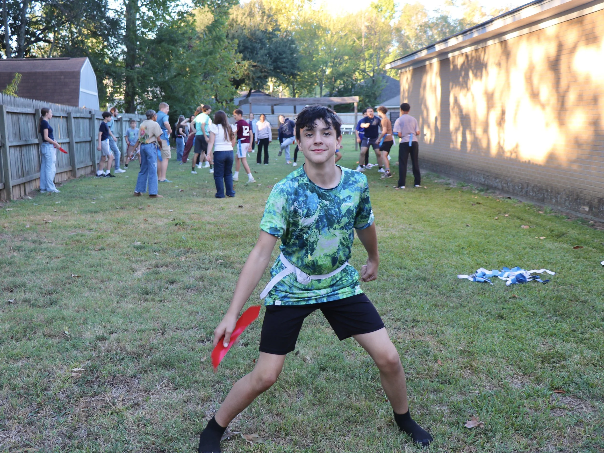 A young boy wearing a colorful shirt and black shorts standing in the Youth House backyard with a group of people playing a game of capture the flag in the background.