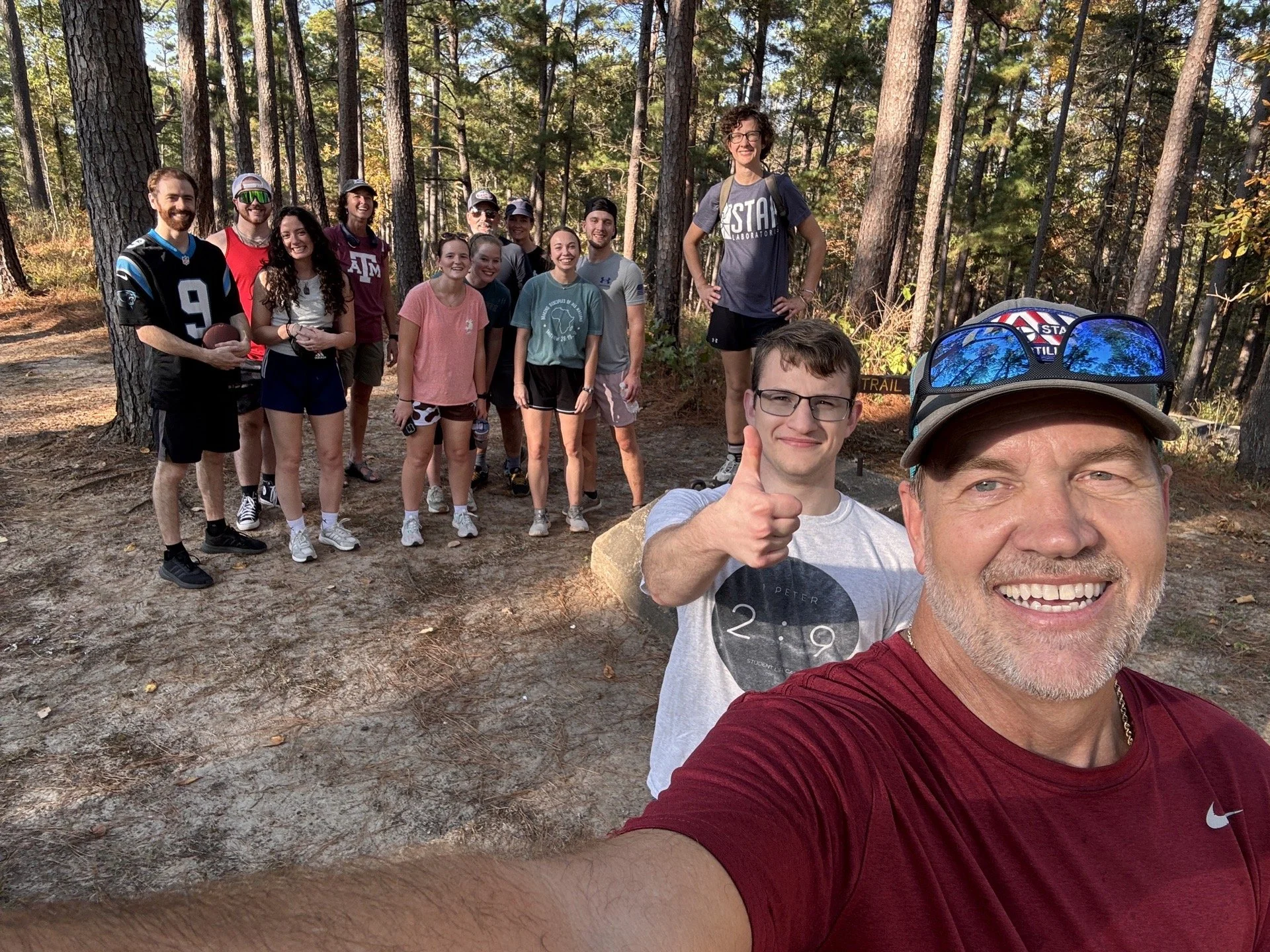 Group of young adults and couples on a wooded trail taking a selfie, with some smiling, giving thumbs-up, and holding a football, surrounded by tall trees and natural outdoor setting.
