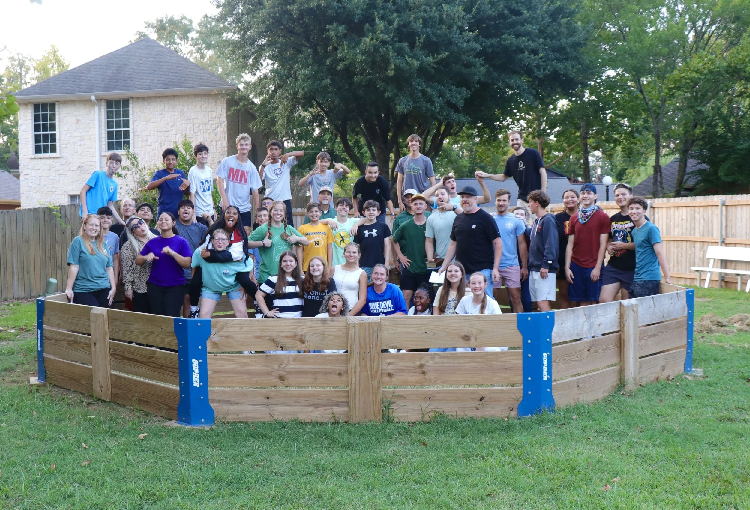 Group of children and adults posing together behind a newly constructed wooden gaga ball pit in the youth house backyard, with trees and houses in the background.