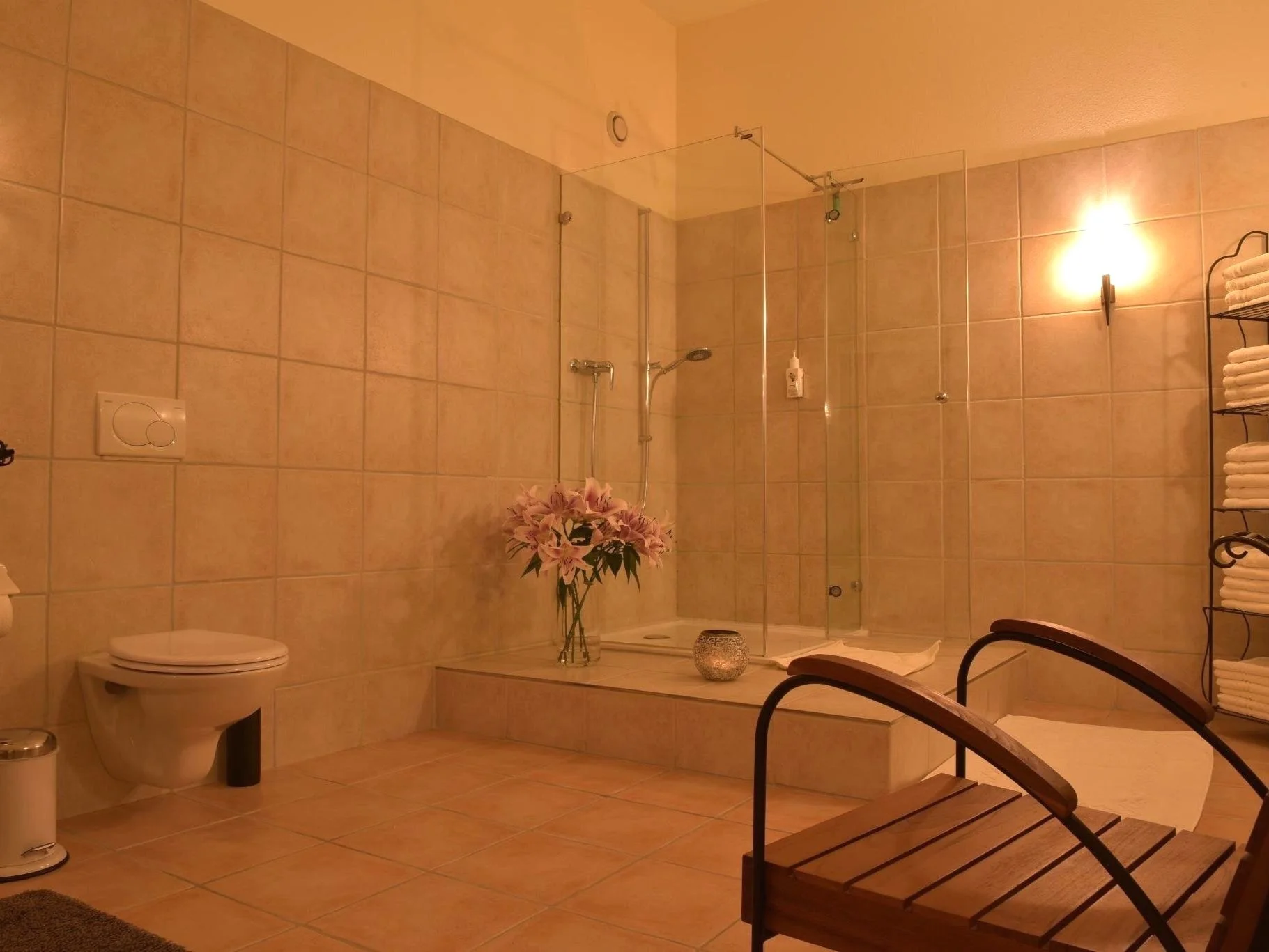 Modernes Badezimmer mit beigen Fliesen, Glasdusche, Waschbecken, Handtüchern auf einem Regal, Blumen in einer Vase und einem Holzstuhl.