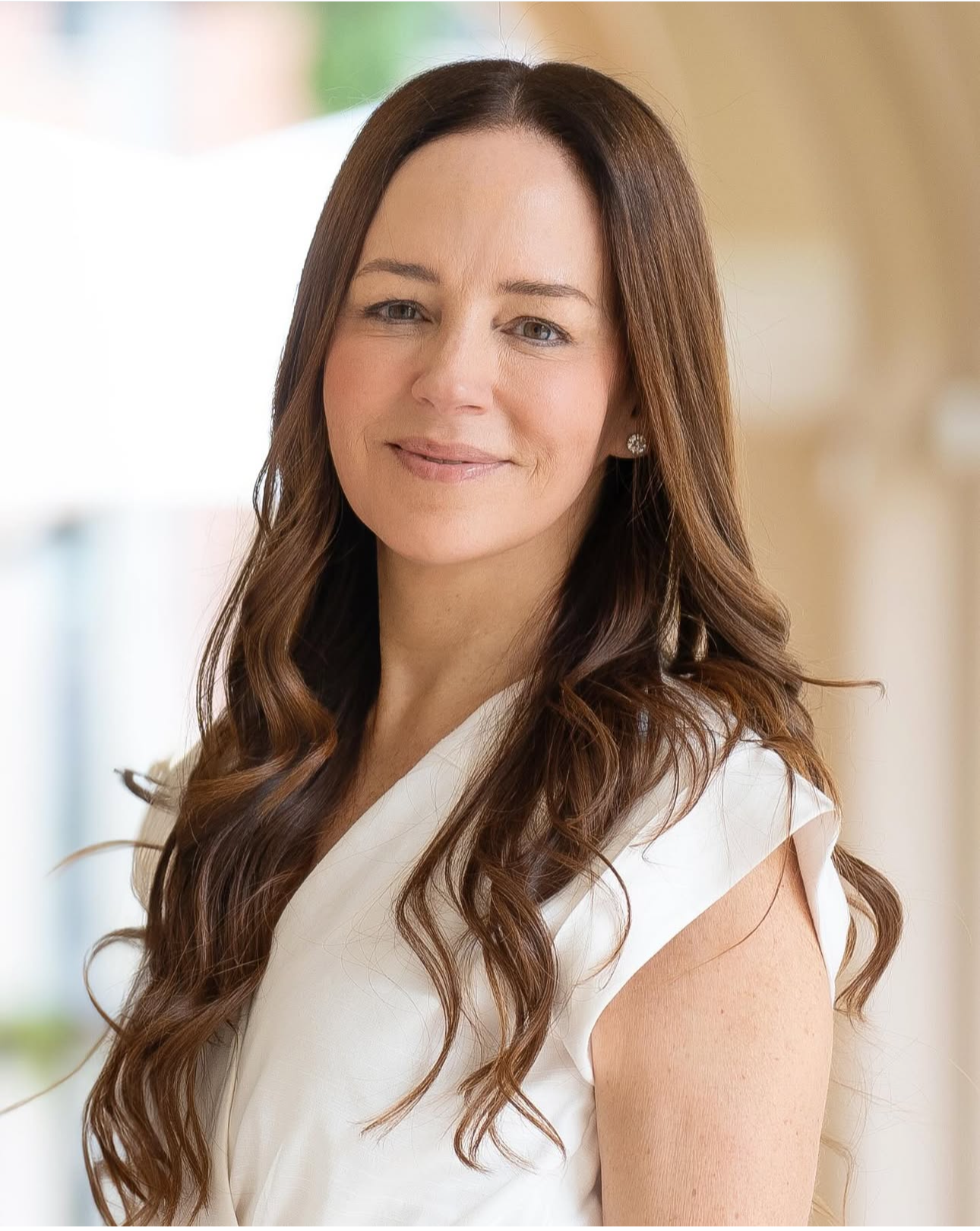 A woman with long, wavy brown hair smiling softly in a light-colored top indoors.