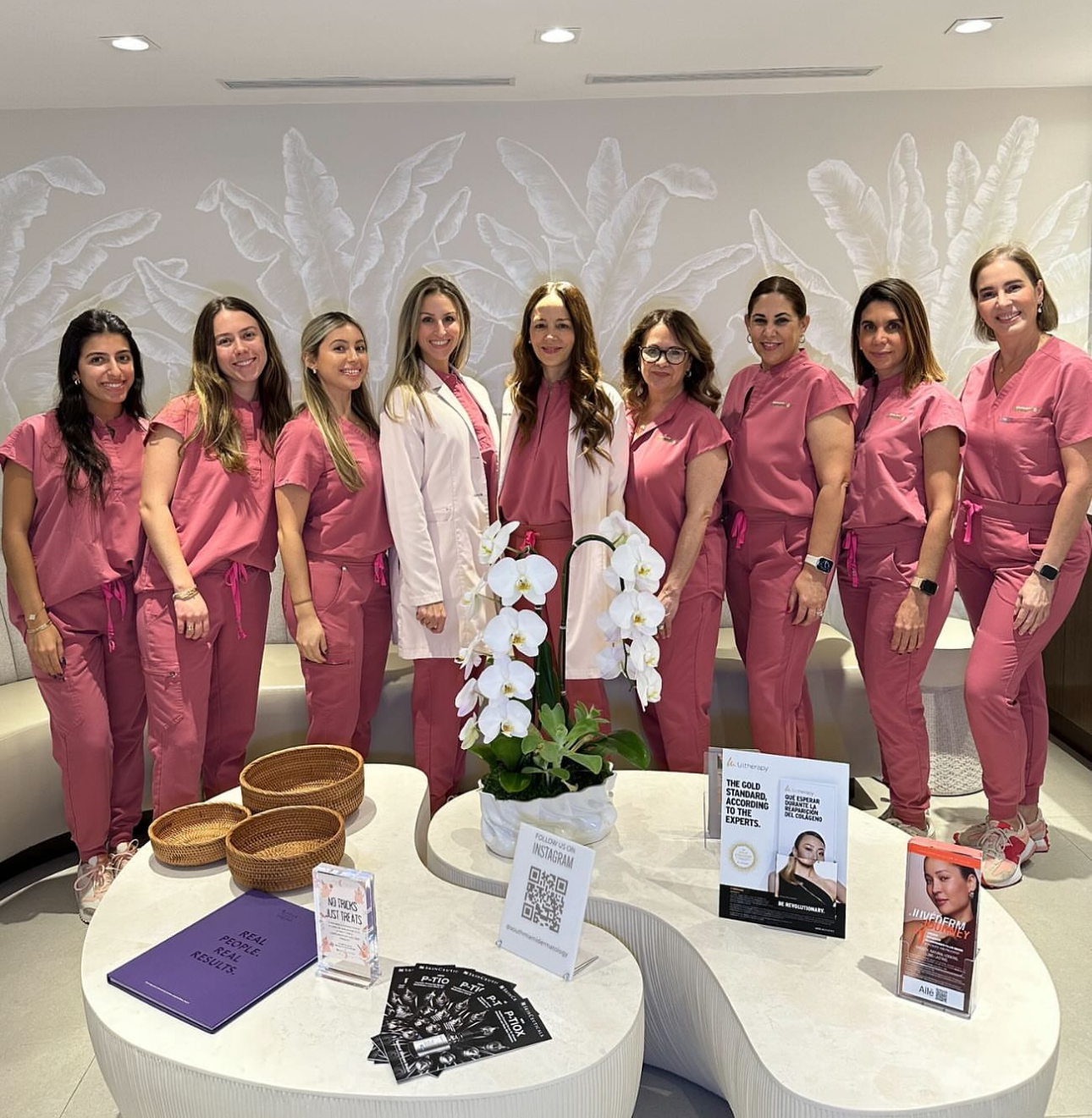 Group of nine women, some in pink medical scrubs and two in white coats, standing indoors in front of a white wall with leaf patterns; a table in front displays orchids, brochures, and signs.