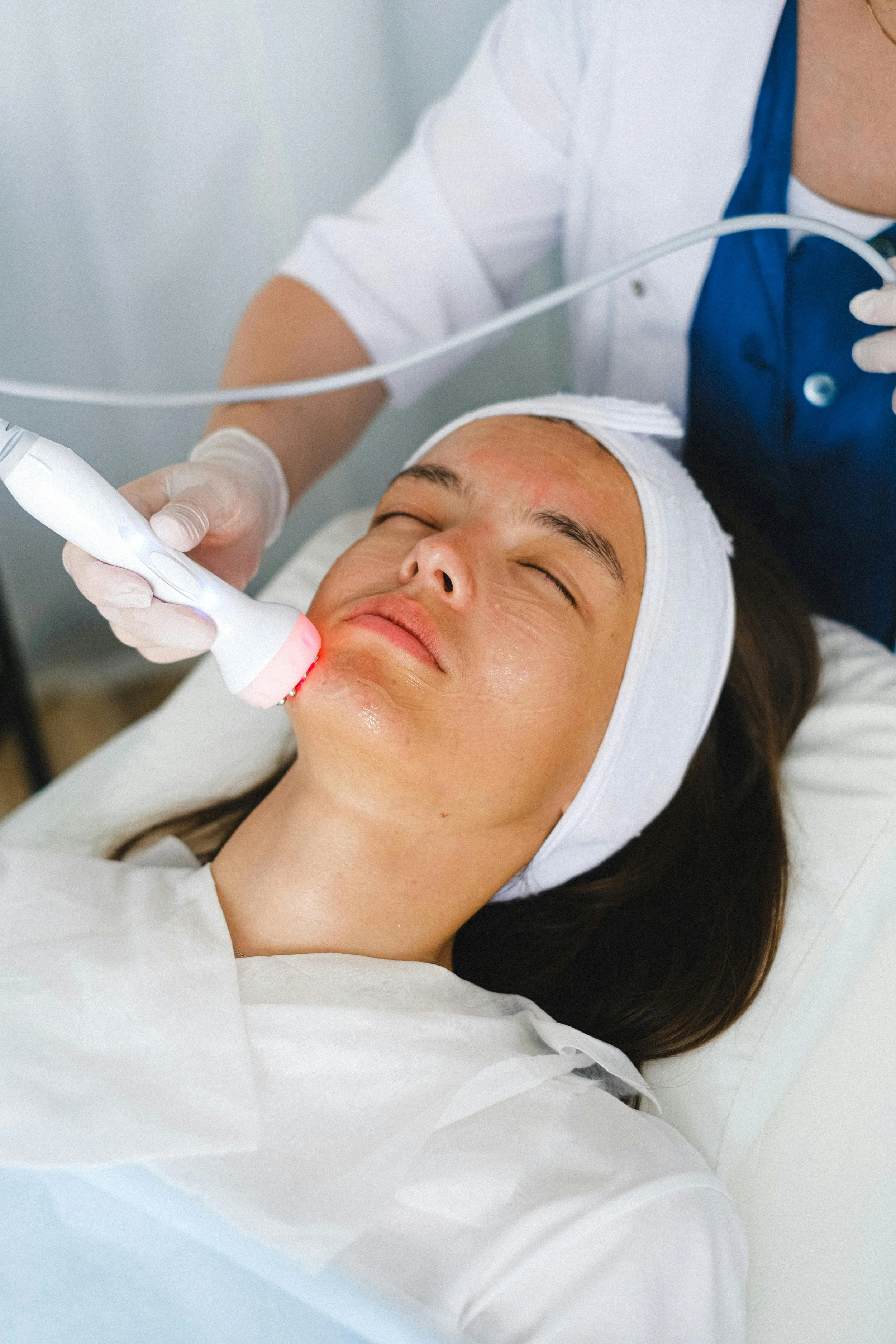 A woman receiving a facial treatment with a handheld device emitting light, lying down with eyes closed, wearing a white headband and gown, in a skincare clinic.