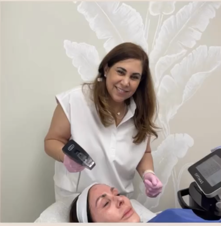 A woman wearing a white outfit and pink gloves smiling while holding a medical device next to a person's face lying on a medical bed.