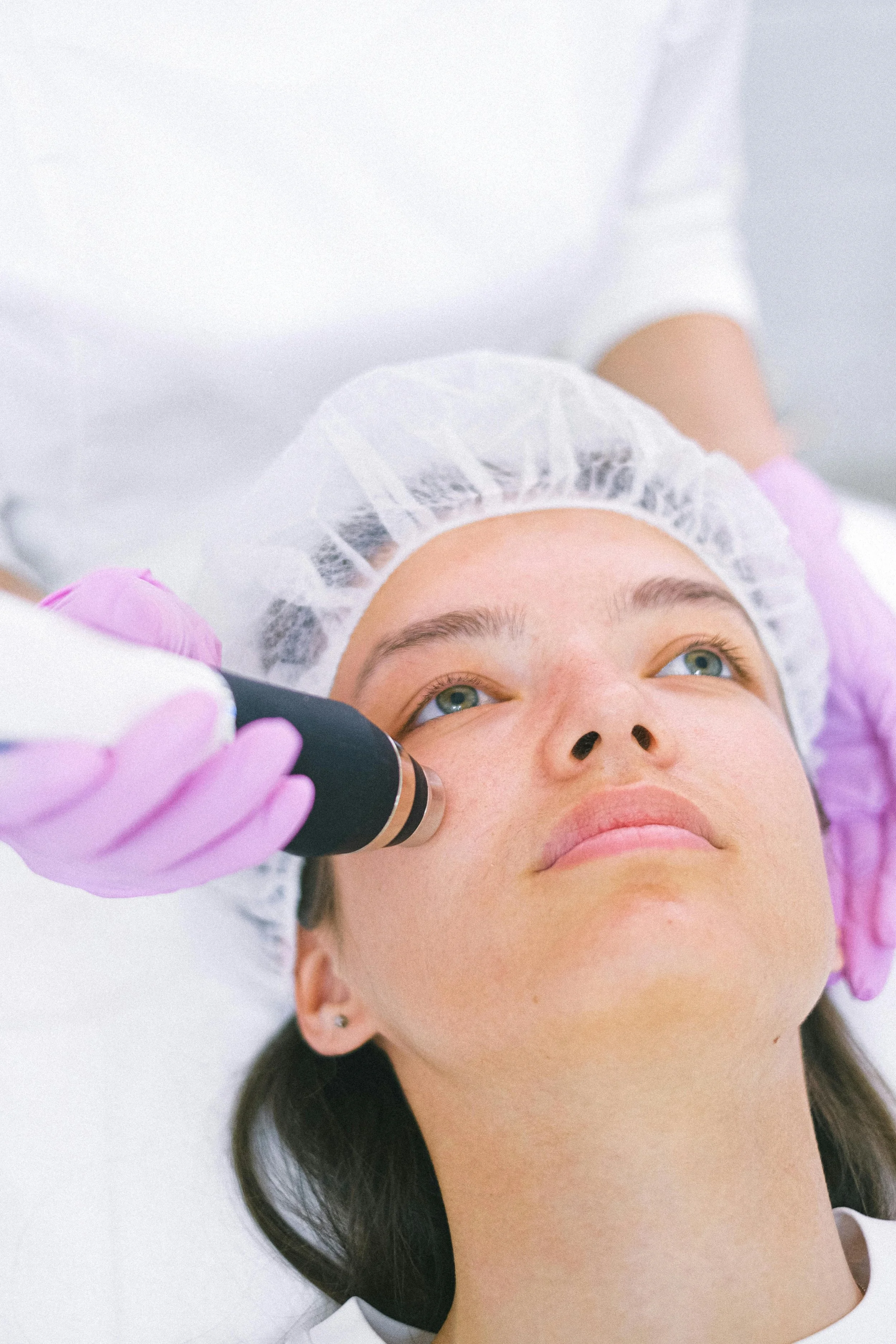 A woman receiving a facial treatment with a handheld device in a clinical setting, wearing a white hair cap and pink gloves.