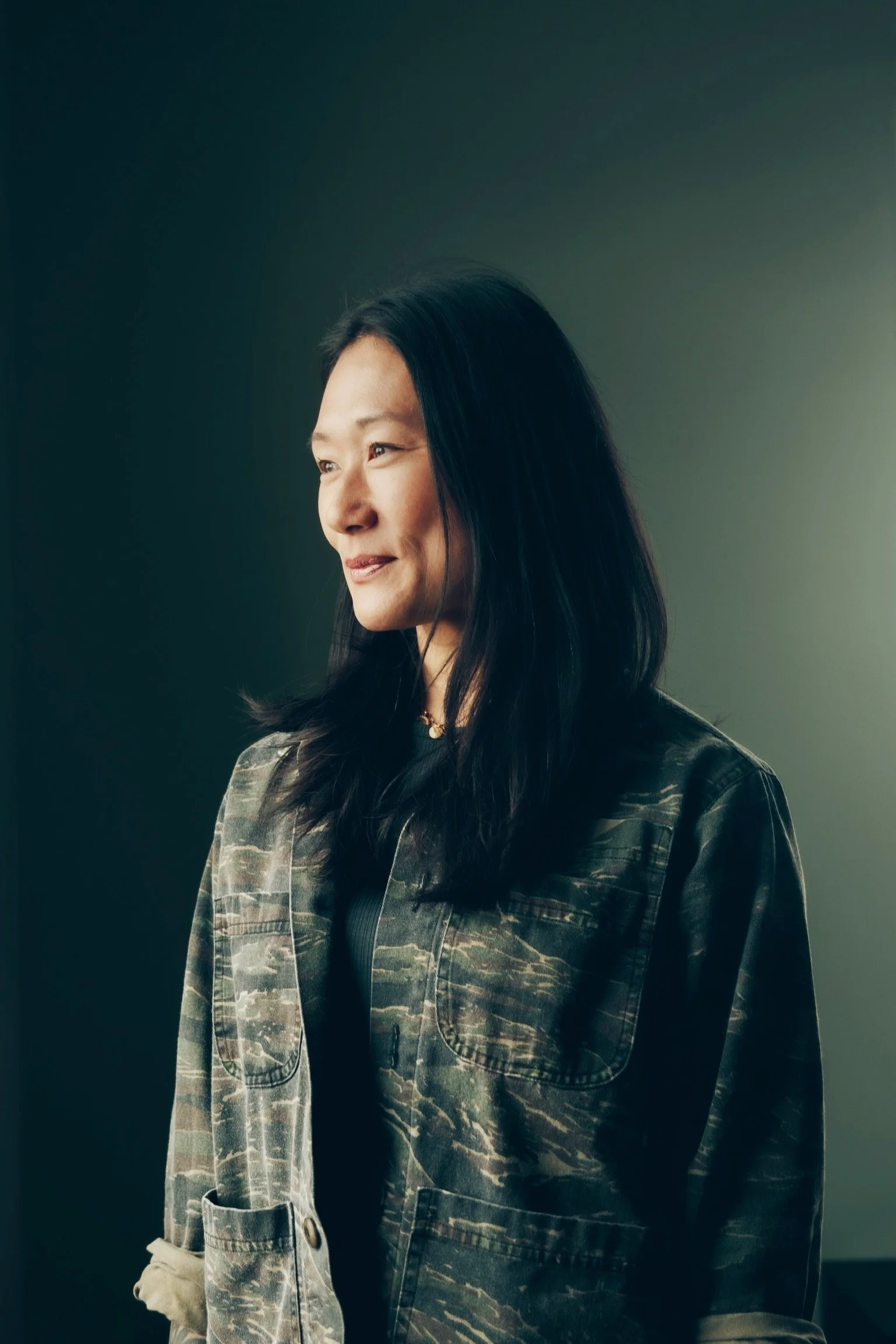 Maggie Carruth O'Connor with long black hair standing in profile against a dark background, wearing a camouflage patterned jacket.
