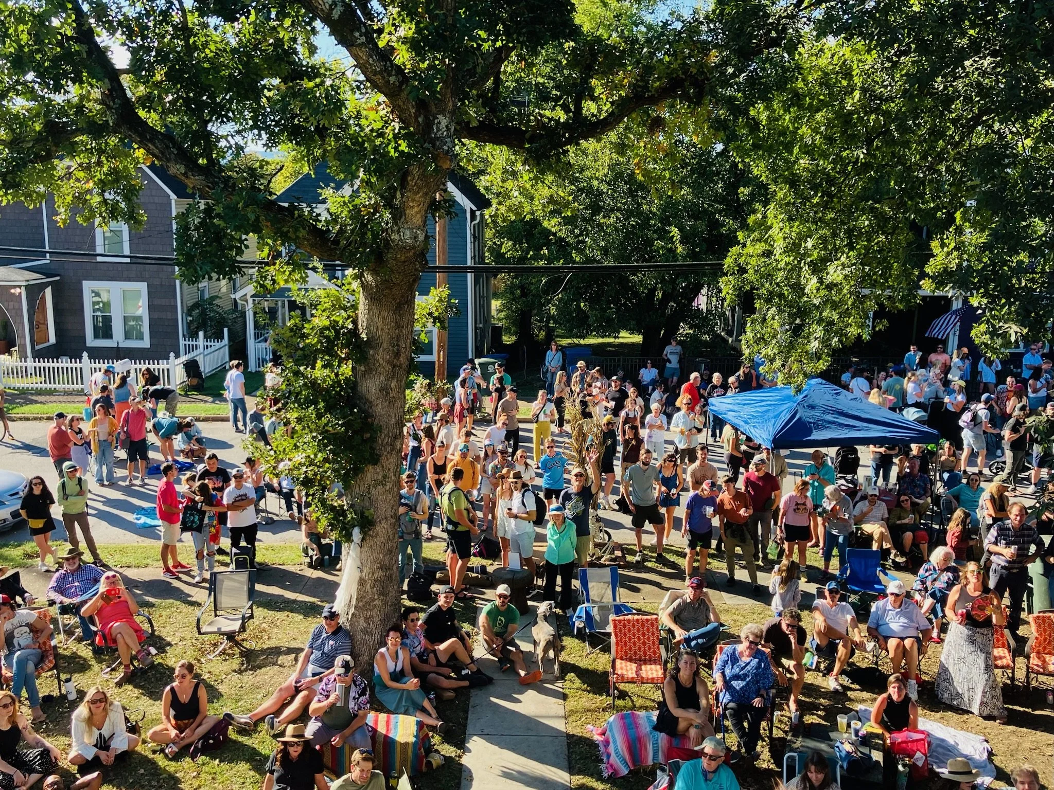 Large crowd of people gathered at Highland Park Porchfest on a sunny day. Some are standing, some are sitting on chairs or blankets. There are trees providing shade, and a blue canopy set up. Houses are visible in the background.