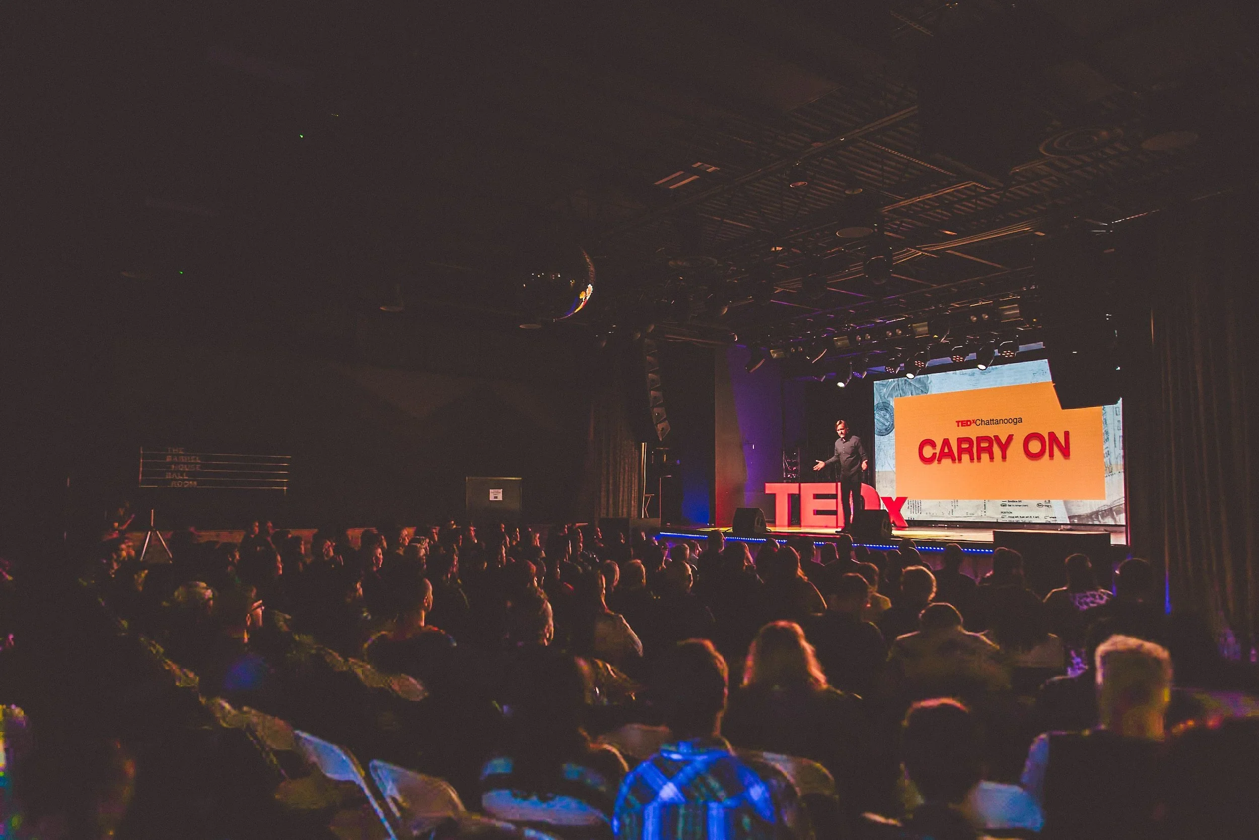 A speaker giving a presentation at a TEDx event on a stage with a large screen behind displaying the words 'Carry On' and 'TEDxChattanooga.' Audience members are seated in a darkened room watching the speaker.